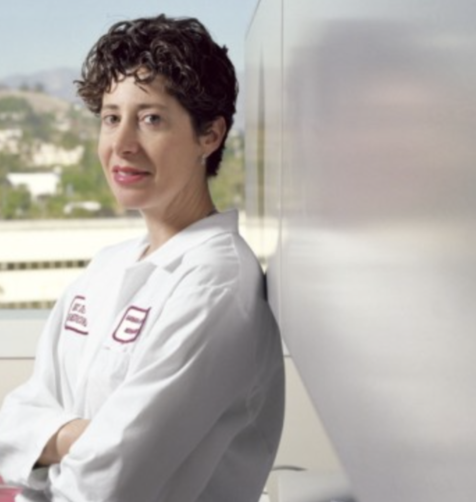 A woman wearing a white lab coat, standing indoors near a window with a cityscape and mountains in the background.