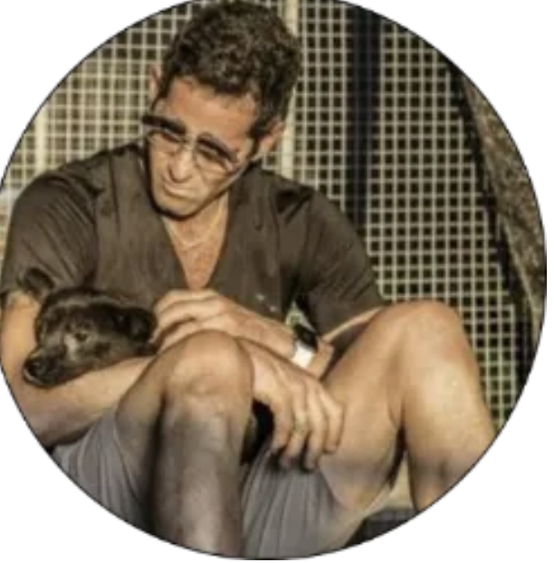 A man sitting with a black dog, holding it gently while sitting against a patterned background.