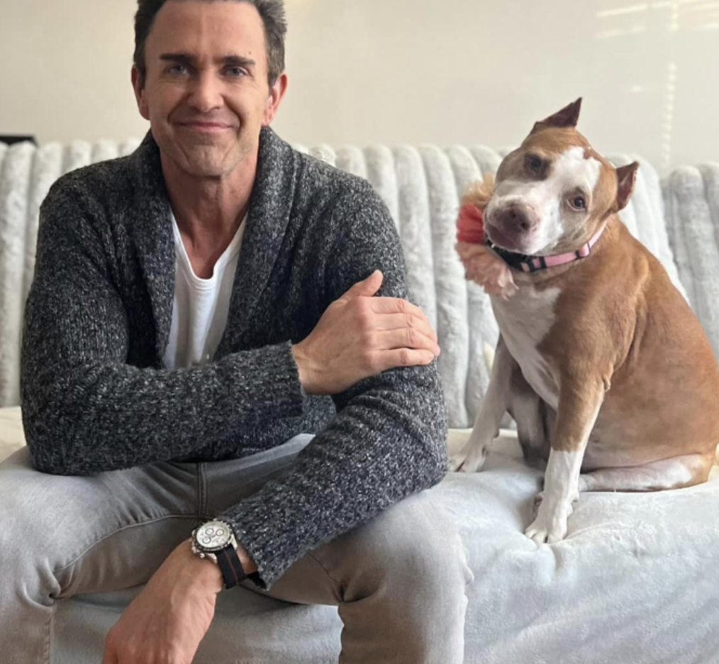 A man with dark hair, wearing a gray cardigan and a white t-shirt, sitting on a light-colored couch, smiling, with an attentive brown and white dog beside him.