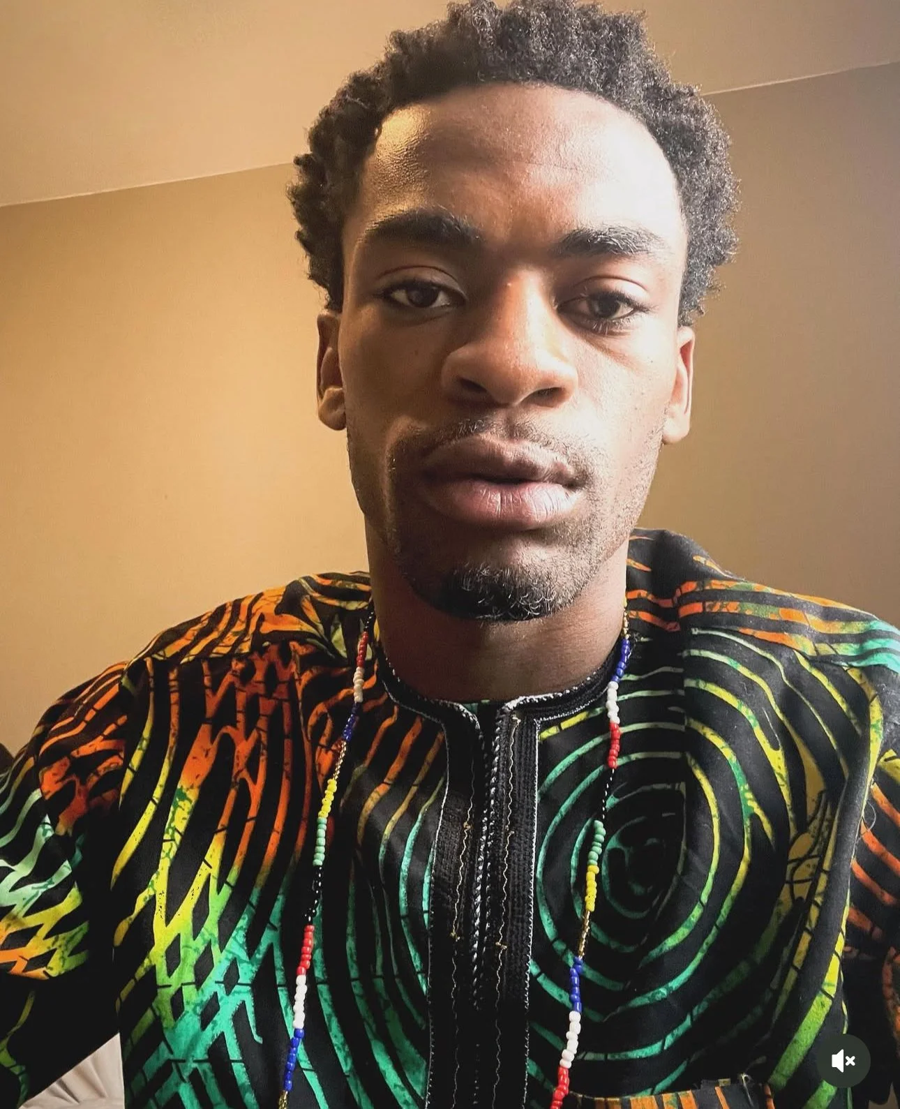 Close-up of a young man with short curly hair wearing a colorful African print shirt with a zipper in the front, and a beaded necklace. He has a neutral expression and is indoors against a plain beige wall.