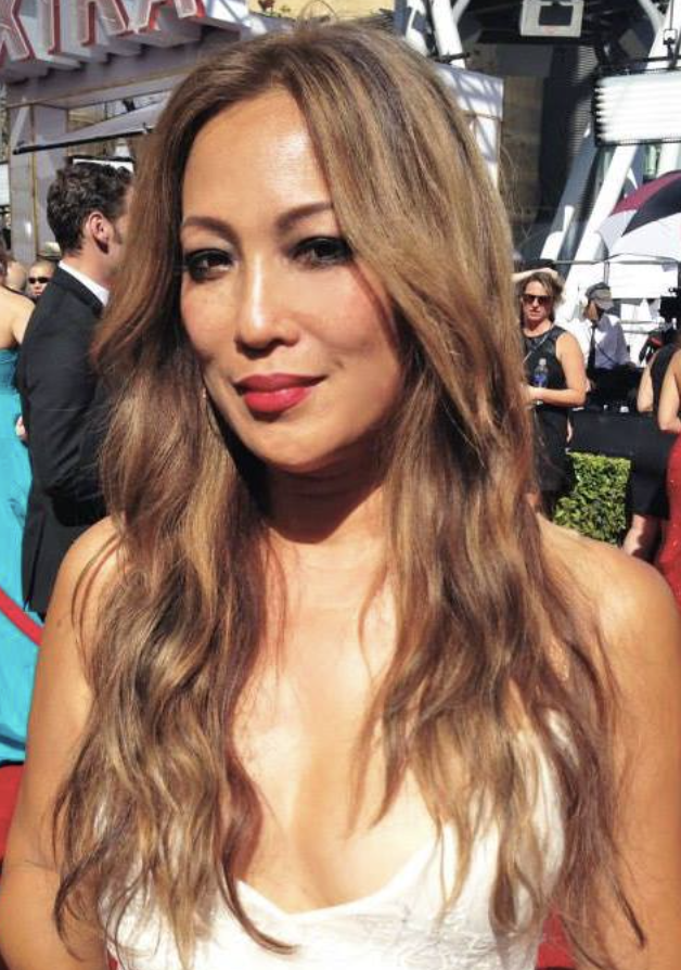 A woman with long, wavy light brown hair and red lipstick at an outdoor event, with people in the background.