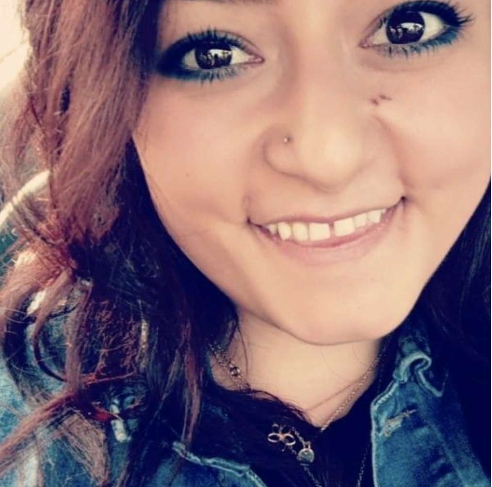 A woman with reddish-brown hair, smiling, wearing a denim jacket with a necklace, and sporting a nose piercing.
