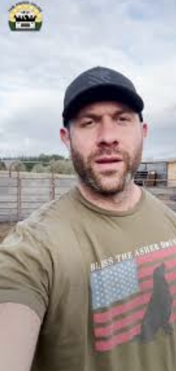 A man in a baseball cap taking a selfie outdoors with a wooden fence and cloudy sky in the background.