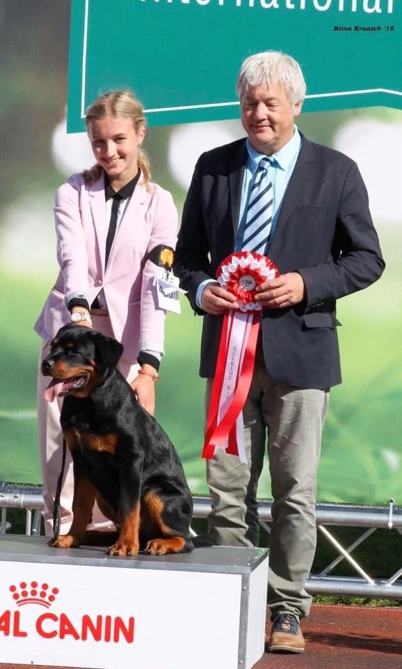 A young woman, a man, and a Rottweiler dog at a dog show. The man is holding a red and white ribbon, and the dog is sitting on a platform with the Royal Canin logo. The background has a blue-green banner.