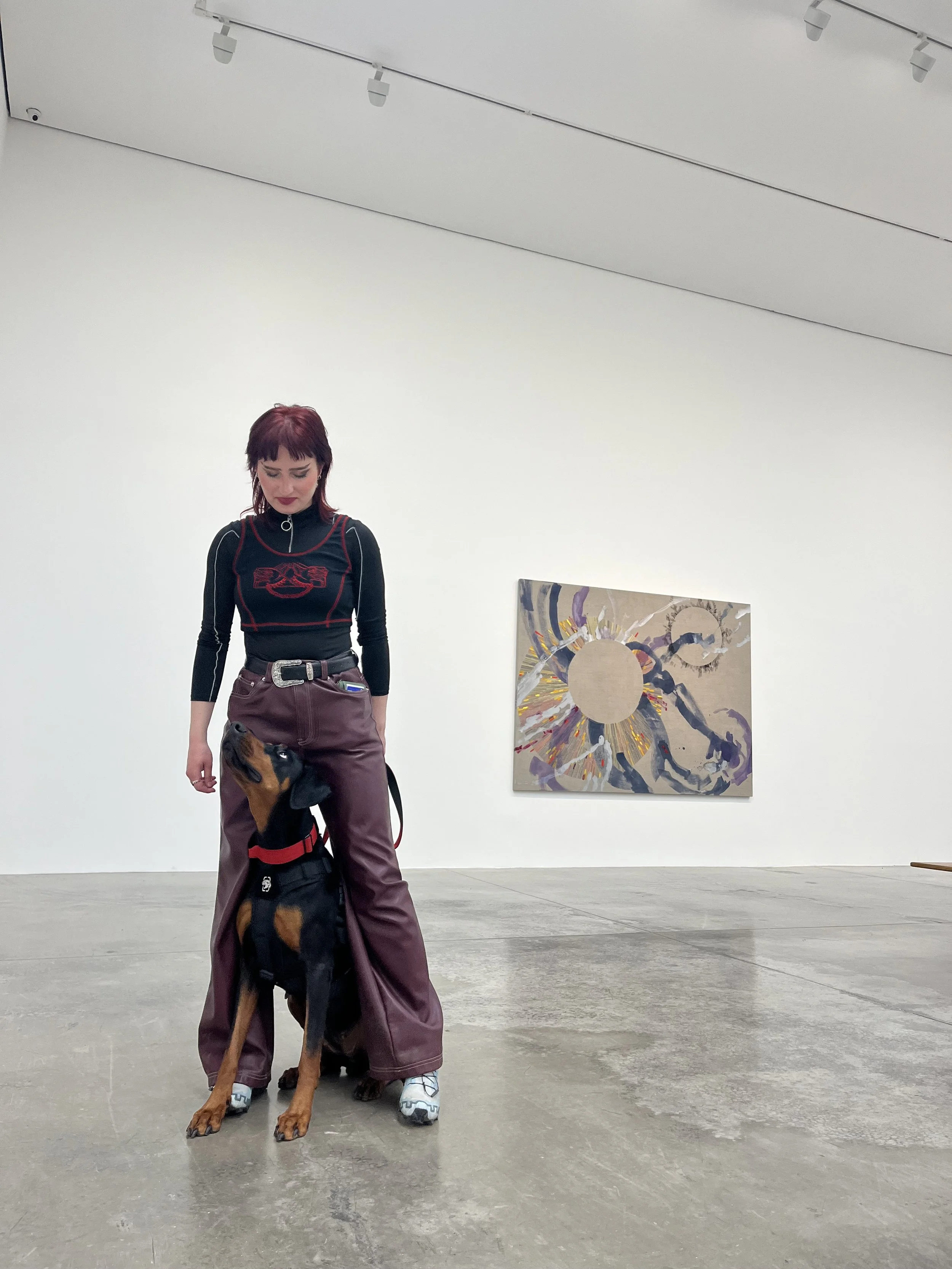 A woman with red hair wearing a black long-sleeve top and maroon wide-leg pants stands on a concrete floor, looking down at a black and brown dog sitting between her legs. The woman is in an art gallery with white walls. There is an abstract painting hanging on the wall behind her.