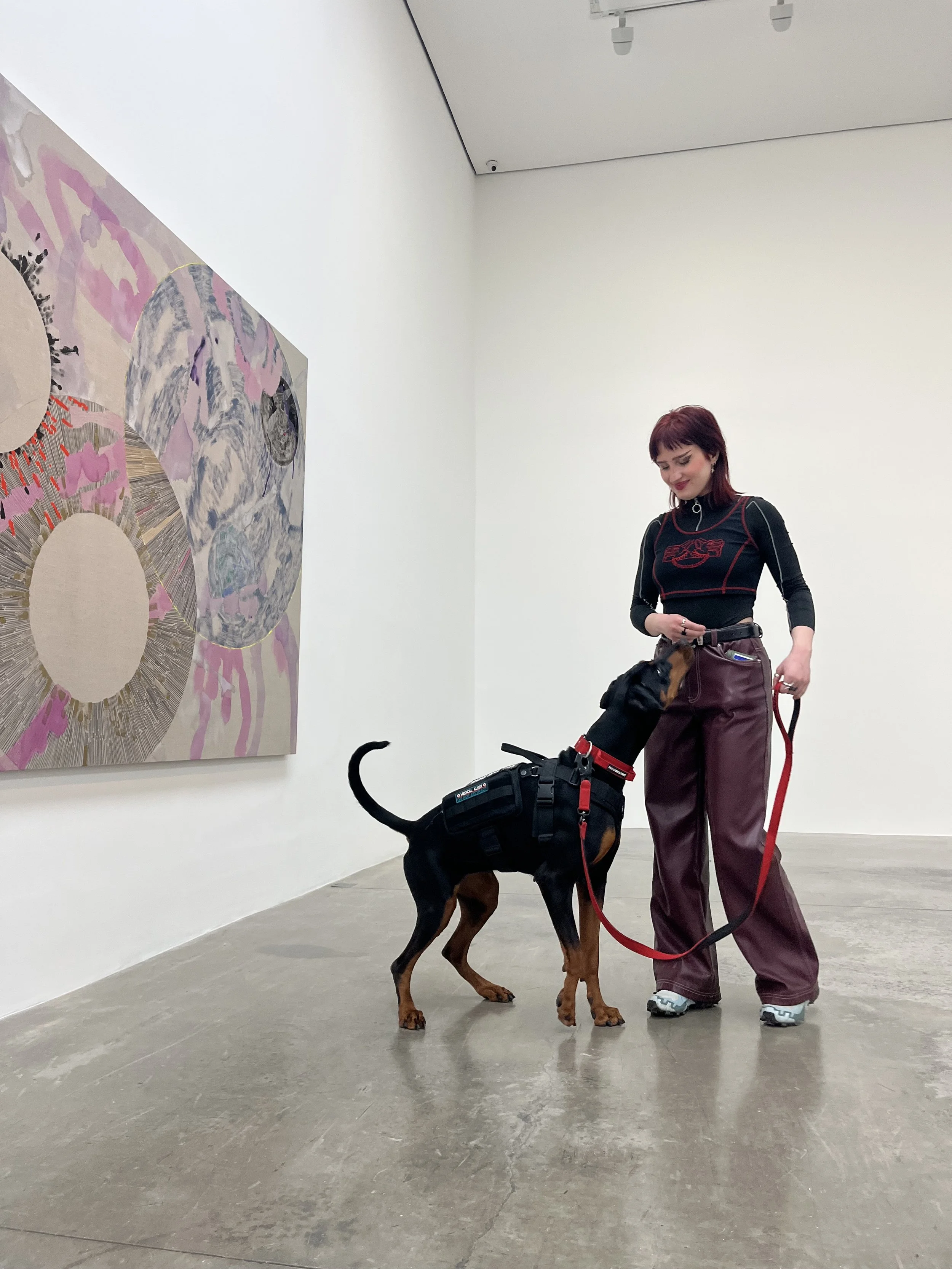 A woman with red hair wearing a black and red shirt and purple pants stands in an art gallery holding a red leash attached to a black and tan guide dog wearing a harness. The woman is smiling while the dog is looking at her. There is an abstract painting on the white wall.