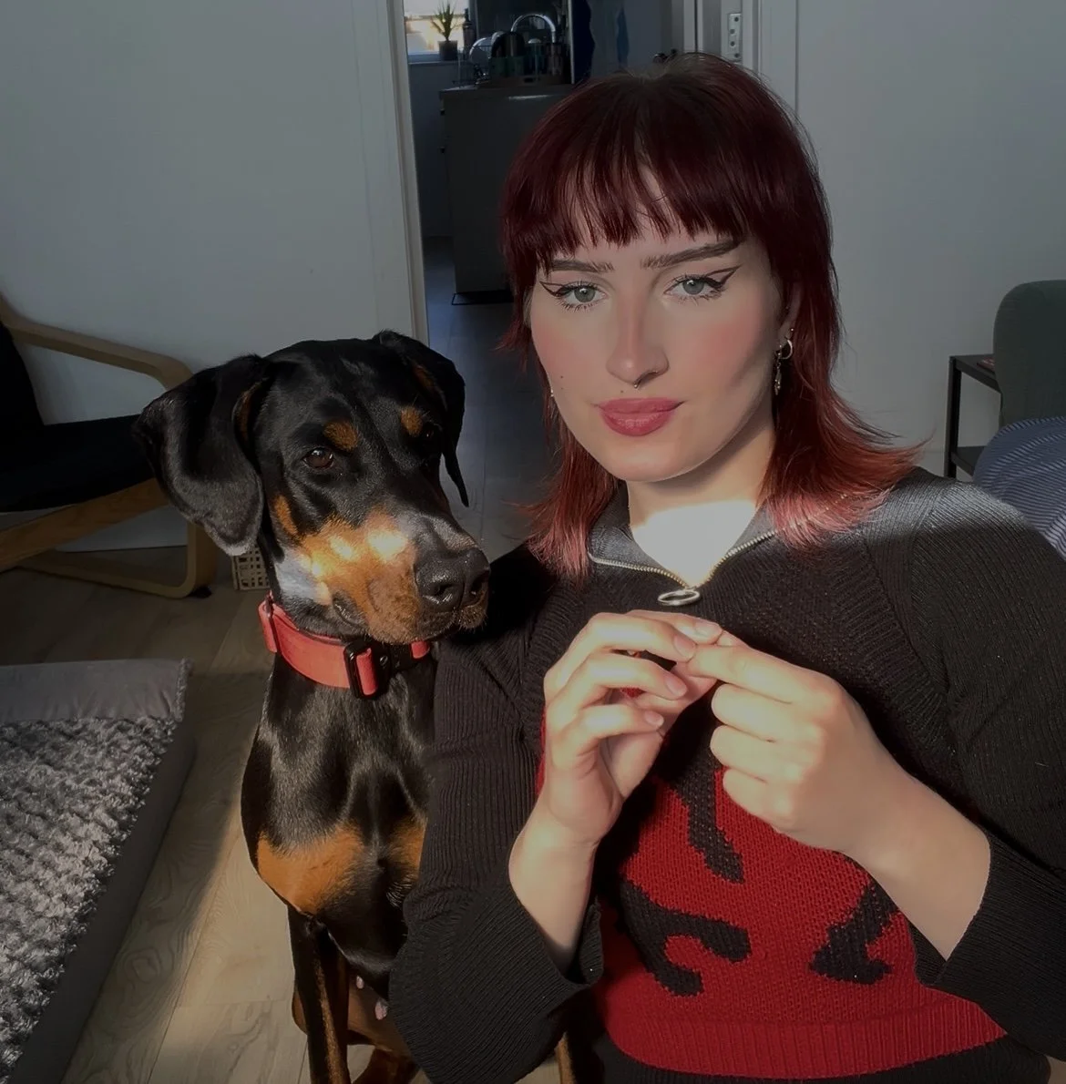 A woman with red hair and winged eyeliner taking a selfie with her dog, a black and tan Doberman, in a living room.