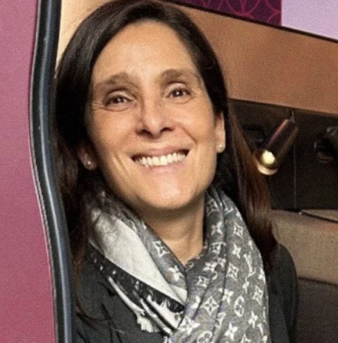 Mulher sorridente com cabelos castanhos, usando escarpa e jaqueta escura, posando em um ambiente interno com fundo em cores rosa e roxa.