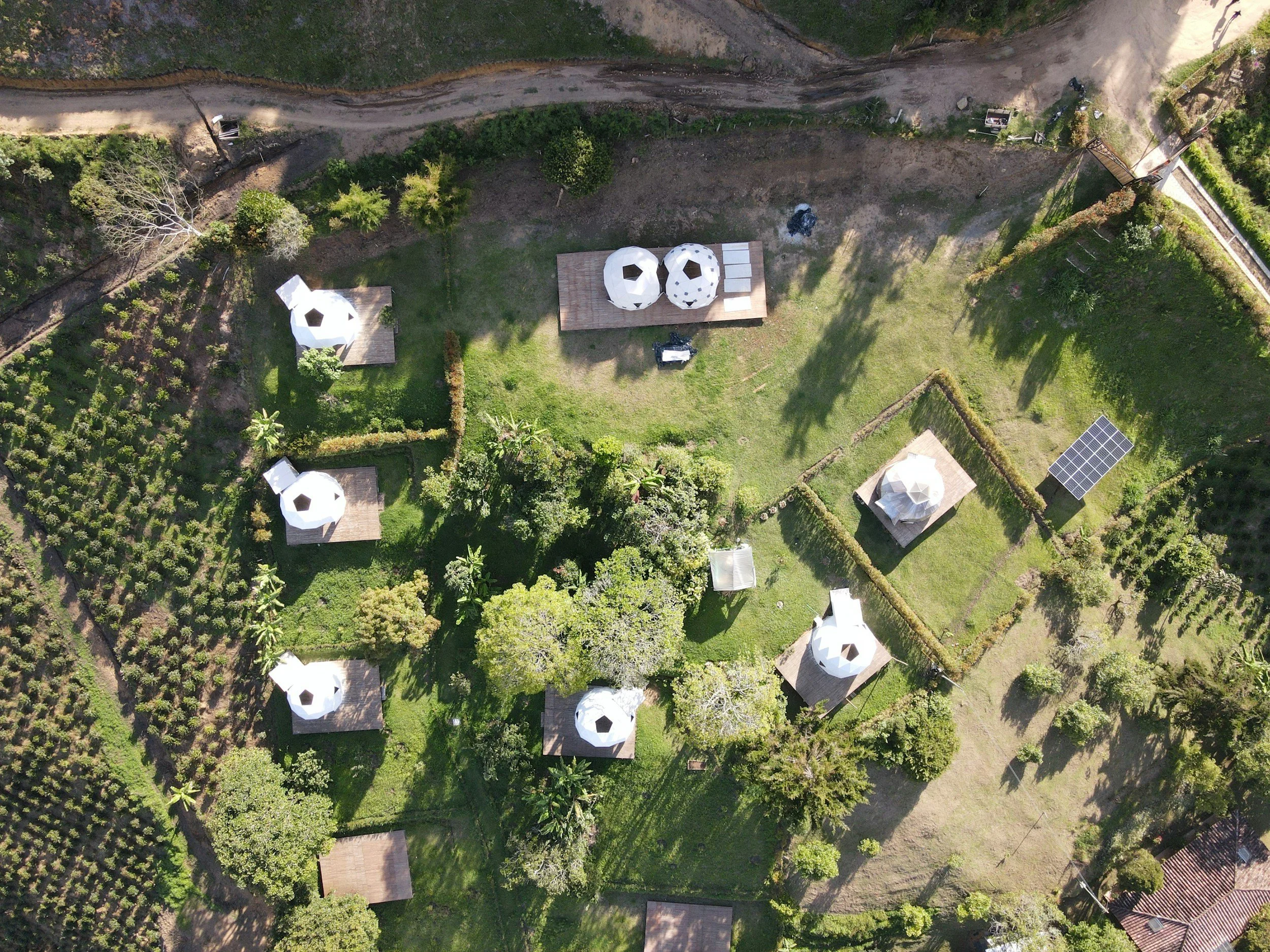 Vista aérea de um terreno com vegetação, árvores, pequeno edifício com telhado de palha, estruturas de vidro com cobertura, painéis solares, e árvores espalhadas, cercado por estrada de terra.