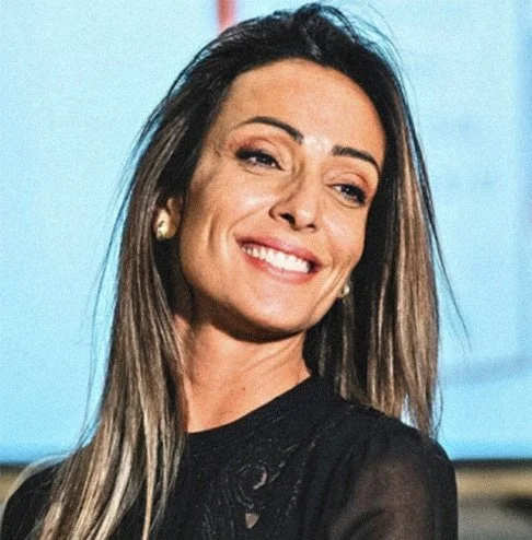 Mulher sorrindo com pele clara, cabelo castanho e liso, usando brincos de argola e roupa preta, em ambiente com fundo azul