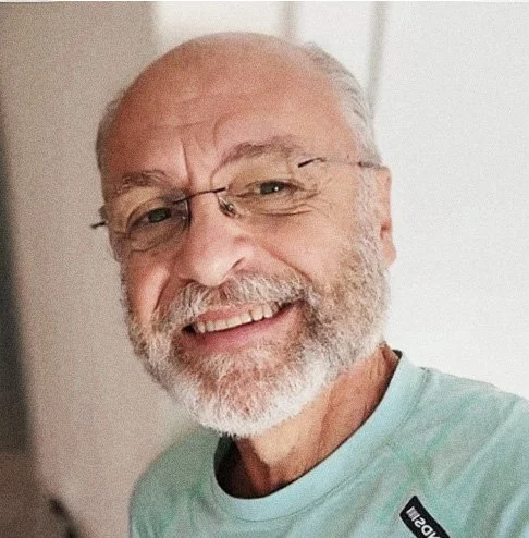 Homem idoso sorrindo, usando óculos, com barba e cabelo grisalho, vestindo uma camiseta verde claro.