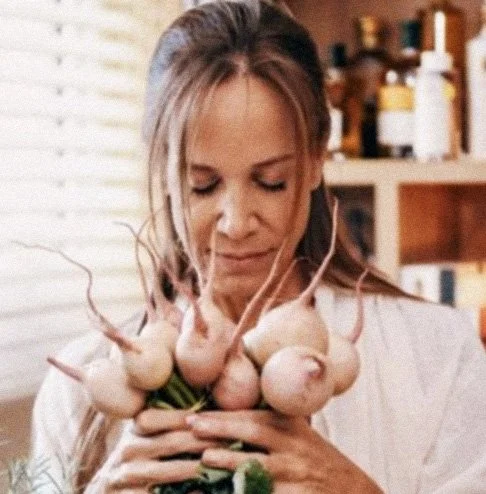 Mulher segurando cebolas na frente do corpo, com uma expressão pensativa. Ambiente de cozinha ao fundo.