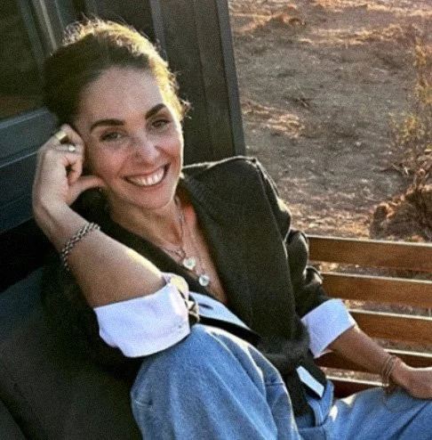 Mulher sorrindo sentada em um banco, com fundo de terra e uma estrutura de vidro, usando jaqueta escura e blusa branca.