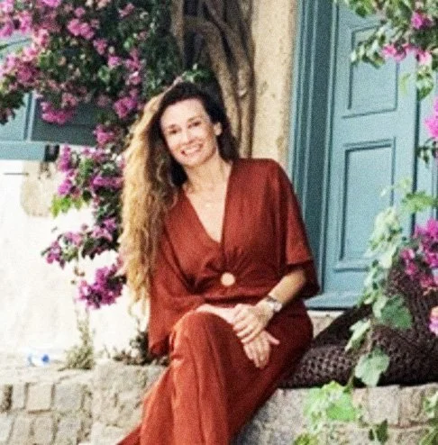 Mulher sorridente de vestido marrom, sentada em um muro de pedra, cercada por flores roxas e paredes de cor azul.