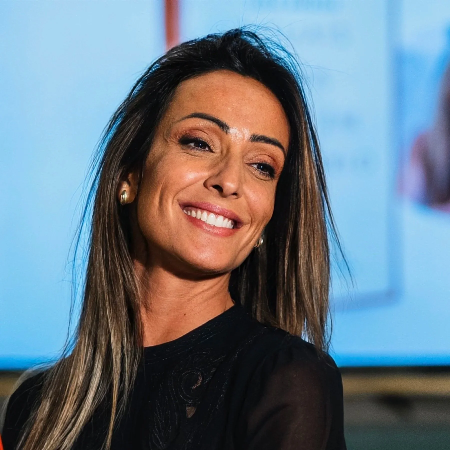 Mulher sorrindo com cabelo castanho claro, usando brincos dourados e fazendo sinal de positivo com a mão. Fundo desfocado