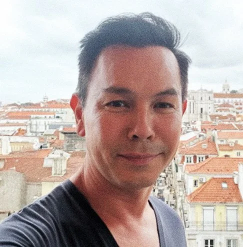 Homem sorridente em primeiro plano com vista de telhados de andares baixos e edifícios históricos ao fundo