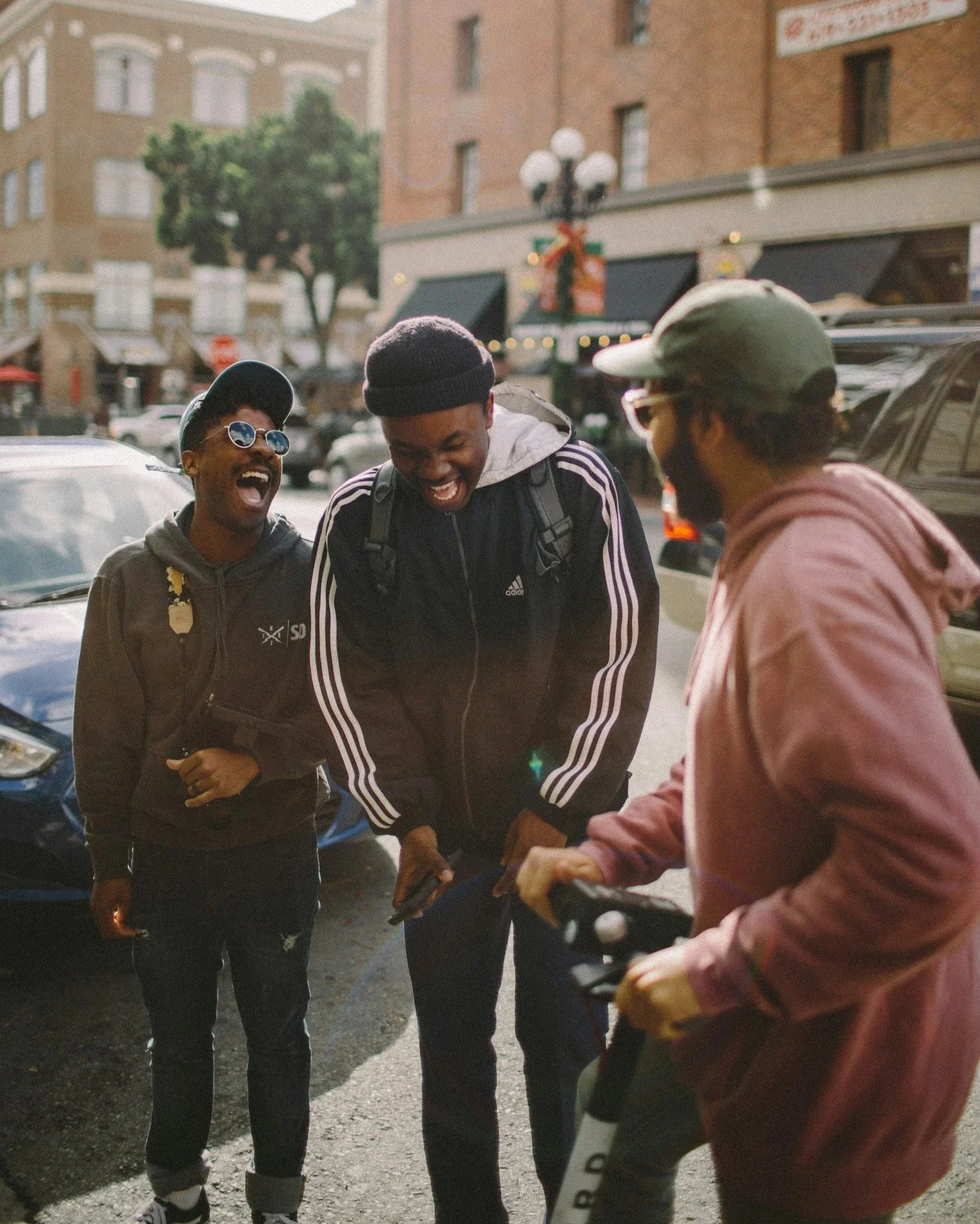Três jovens homens compartilhando uma risada na rua, com prédios e carros ao fundo, durante o dia.
