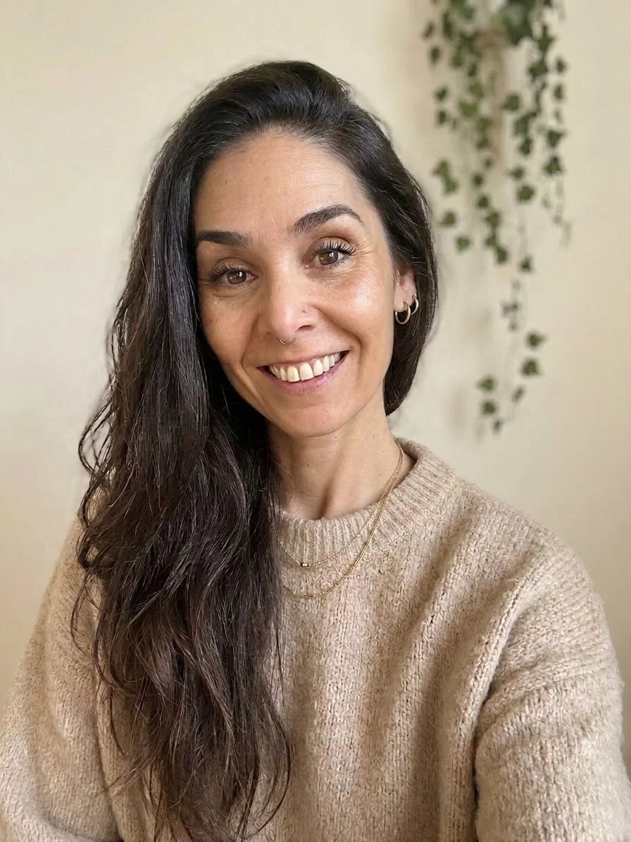 Mulher com cabelo preso, usando blusa de alças cinza, olhando para o lado, com fundo de madeira.