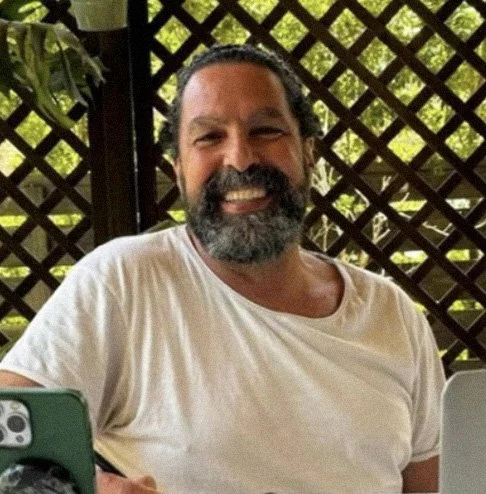 Homem de cabelo escuro e barba grisalha sorridente, sentado ao ar livre com estrutura de treliça de madeira ao fundo e vegetação ao redor.