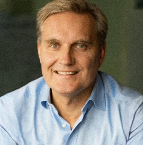 Retrato de um homem com cabelo cinza, usando uma camisa azul clara, sorrindo, com fundo desfocado