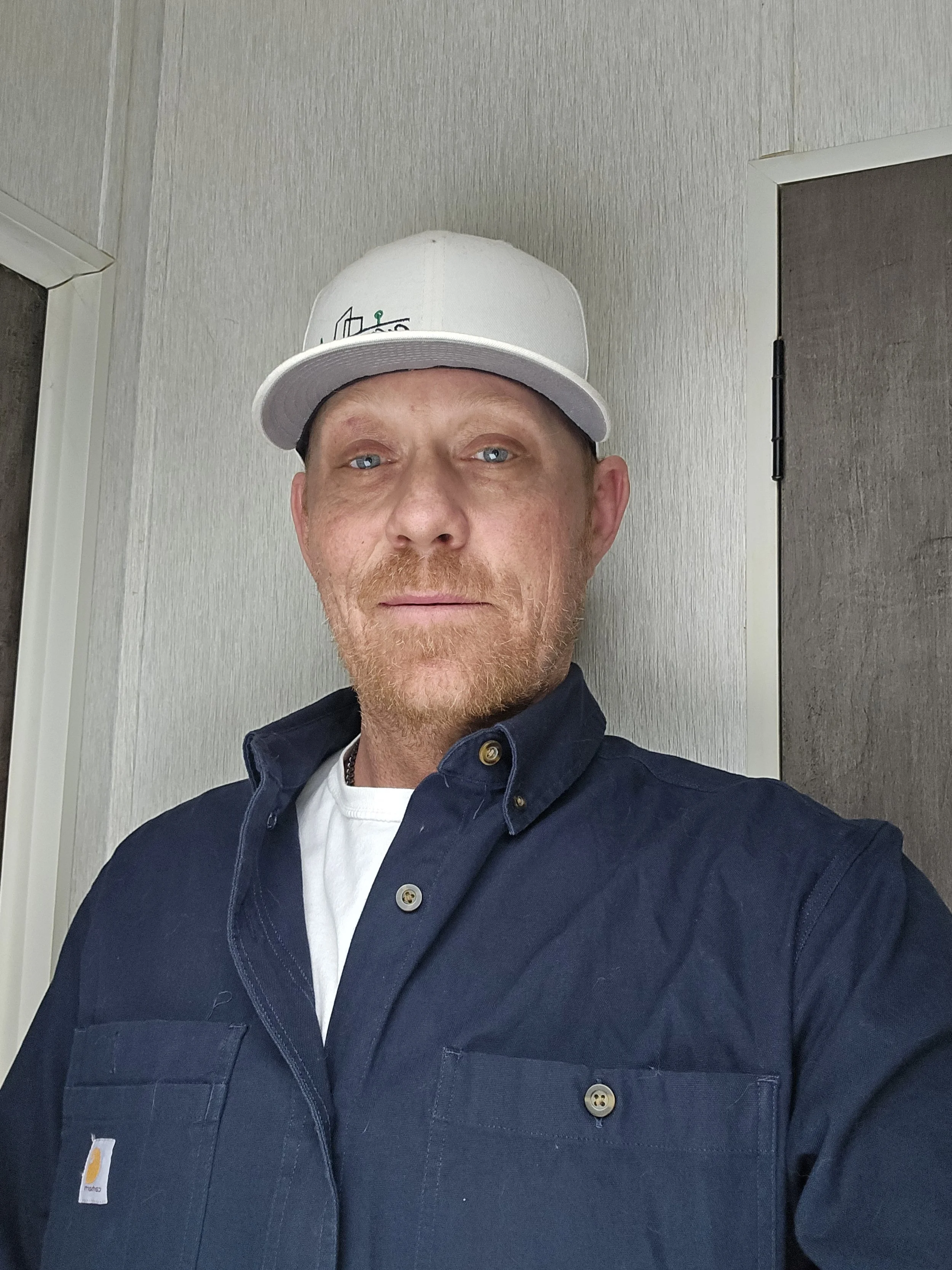 A man with light skin, red hair, and a reddish beard wearing a white cap, a white t-shirt, and a dark blue button-down shirt, standing indoors against a light-colored wall and a closed door.