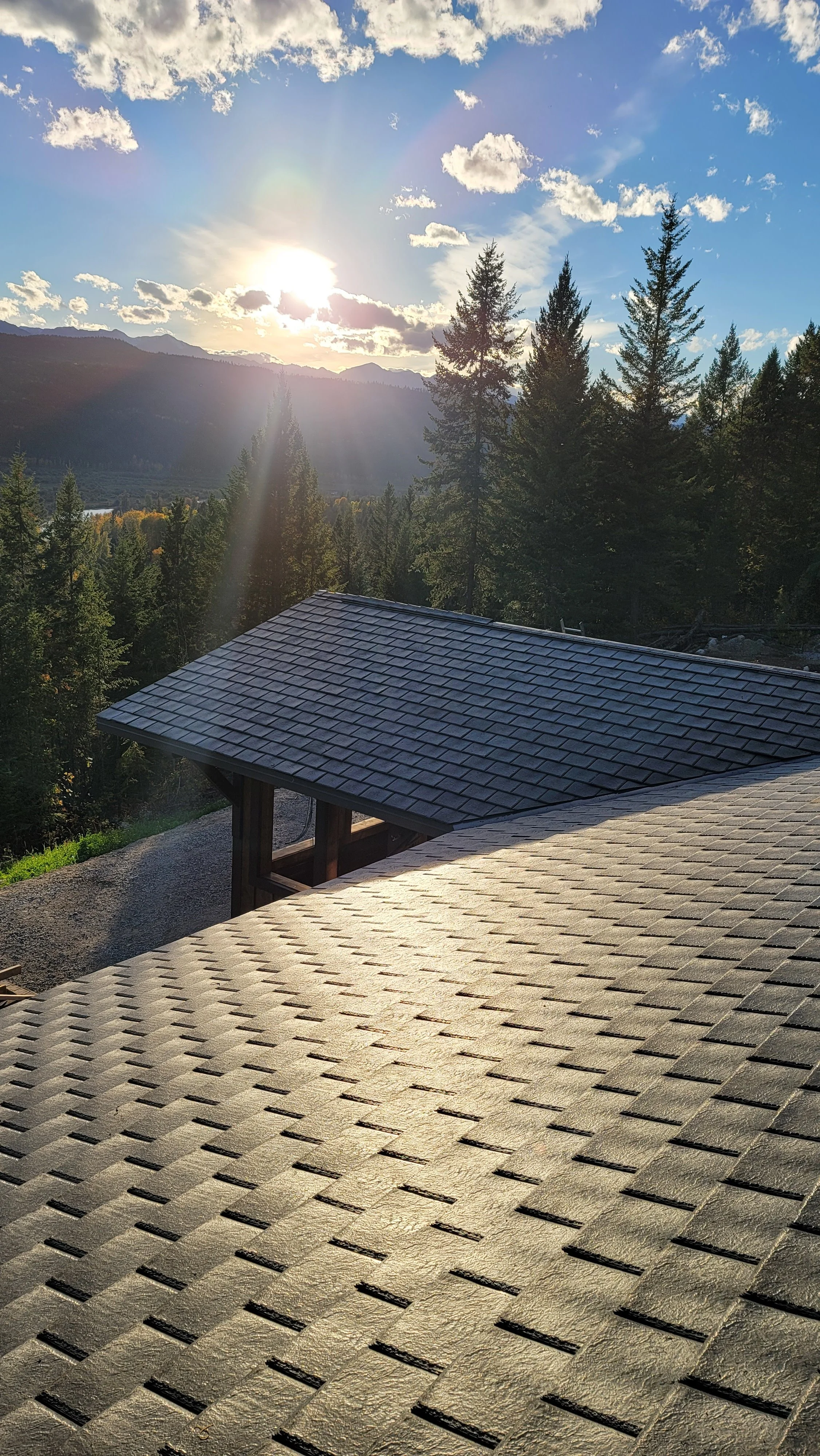 Sunset over a mountainous landscape with tall pine trees, viewed from a roof with asphalt shingles.