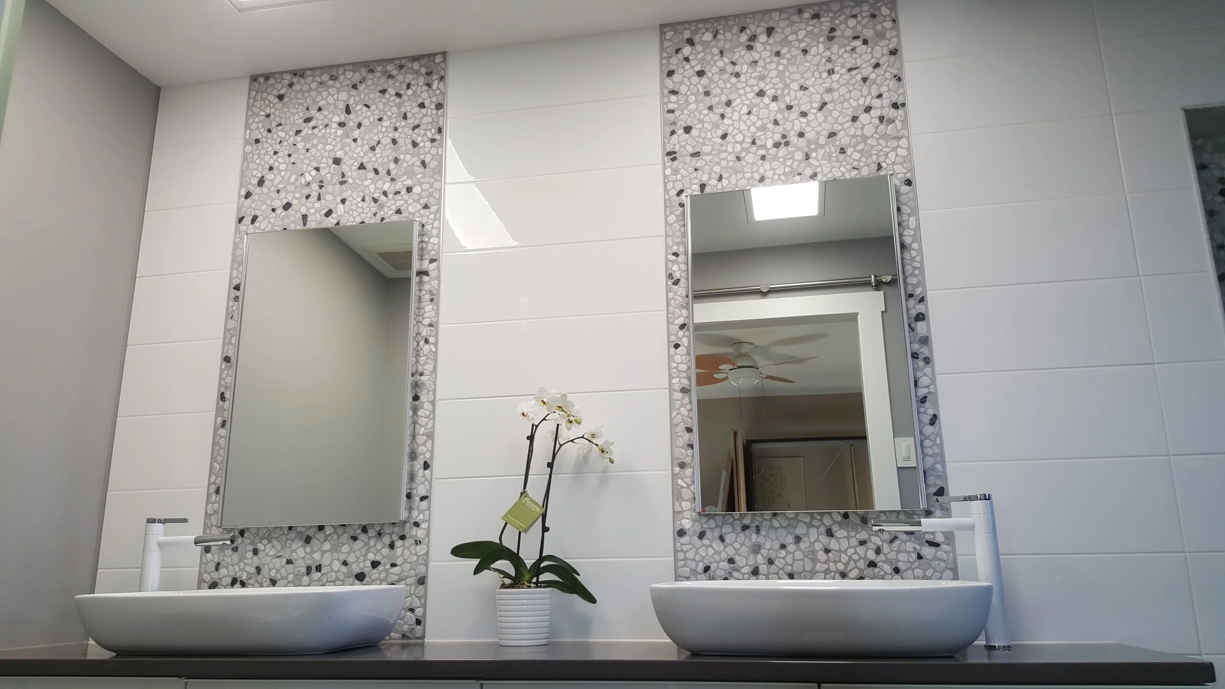 Bathroom with two white vessel sinks, each with a modern faucet, set on a dark countertop. Two large, rectangular mirrors with mosaic tile borders are mounted on a white tiled wall. A potted white orchid with a green tag is placed between the sinks. Reflections in the mirrors show more of the bathroom and a ceiling fan in an adjoining room.