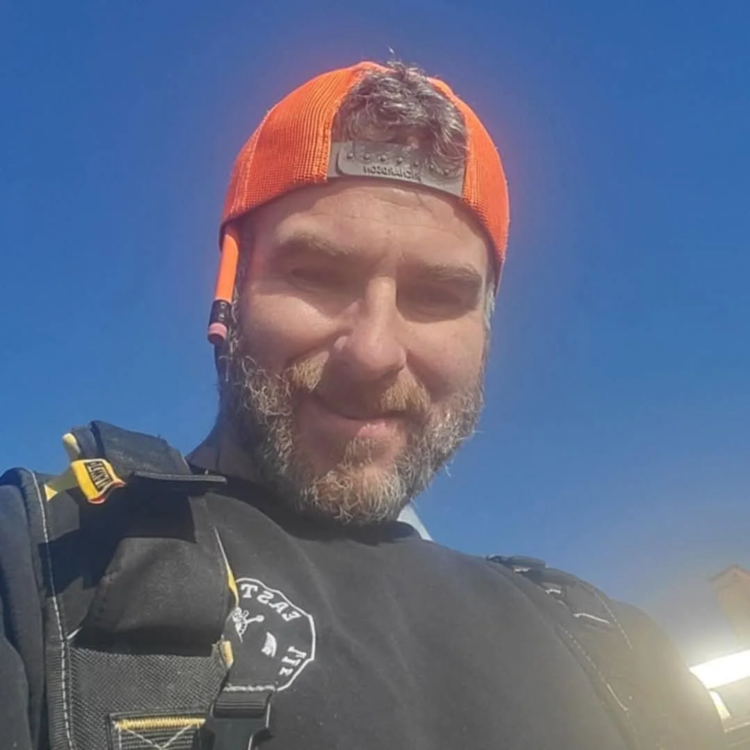 A man wearing an orange cap, black shirt, and a backpack, smiling outdoors against a clear blue sky.