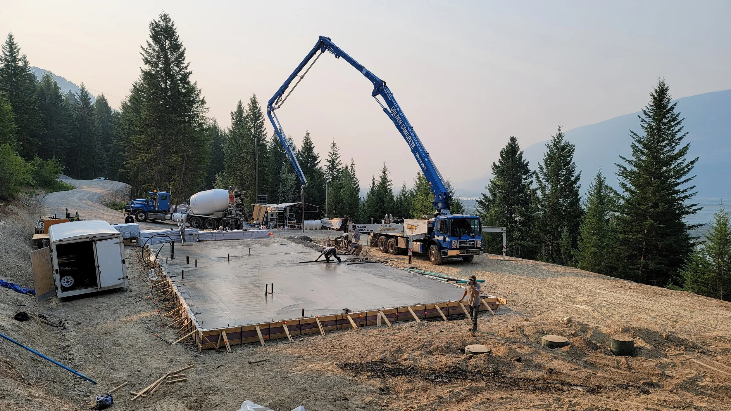 Construction workers pouring concrete for a building foundation on a hillside surrounded by trees, with a cement mixer truck and a concrete pump truck in operation.