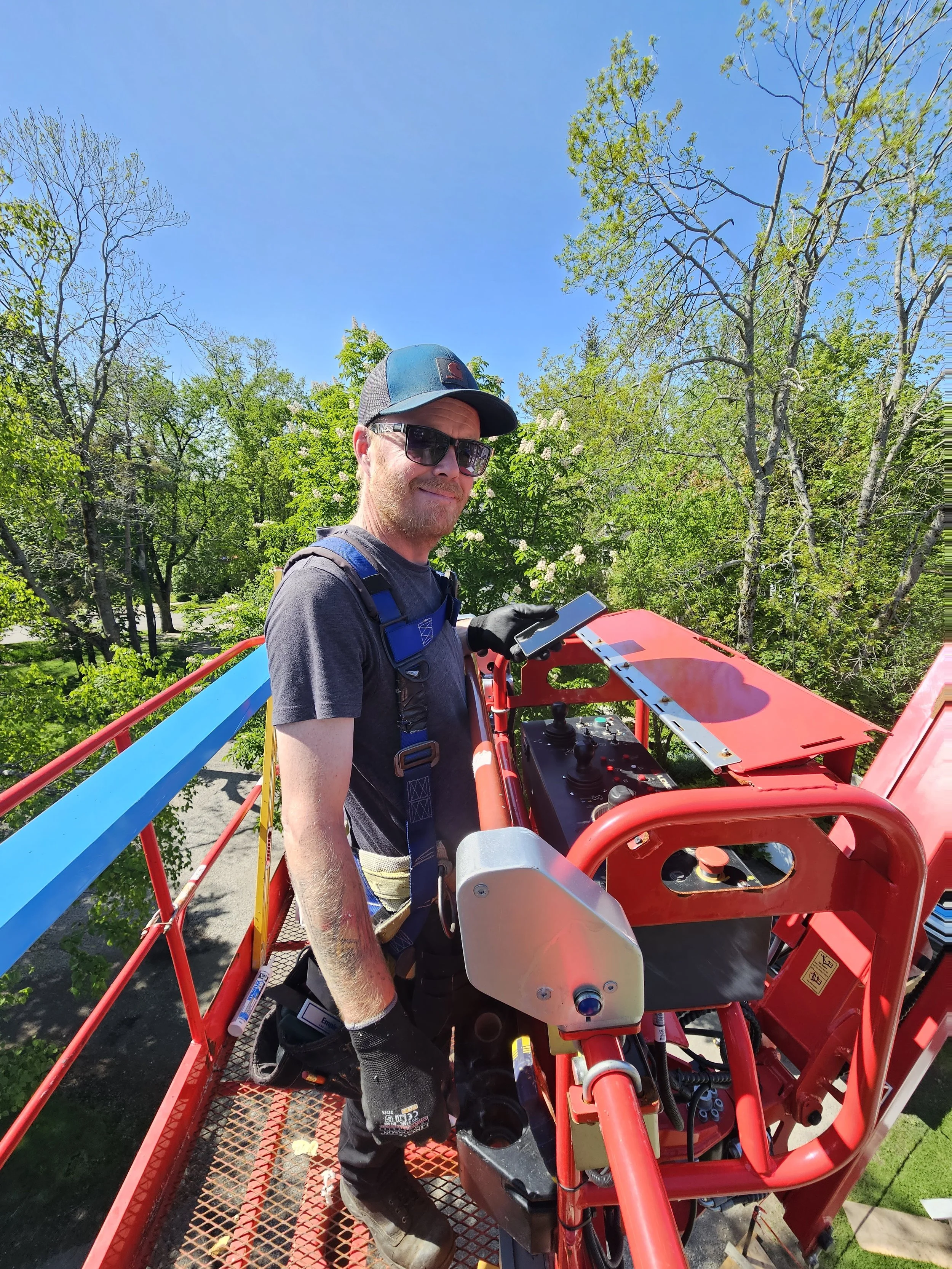 Man standing on a cherry picker lift platform, wearing sunglasses, a baseball cap, gloves, and a harness, operating machinery amidst green trees under a bright blue sky.