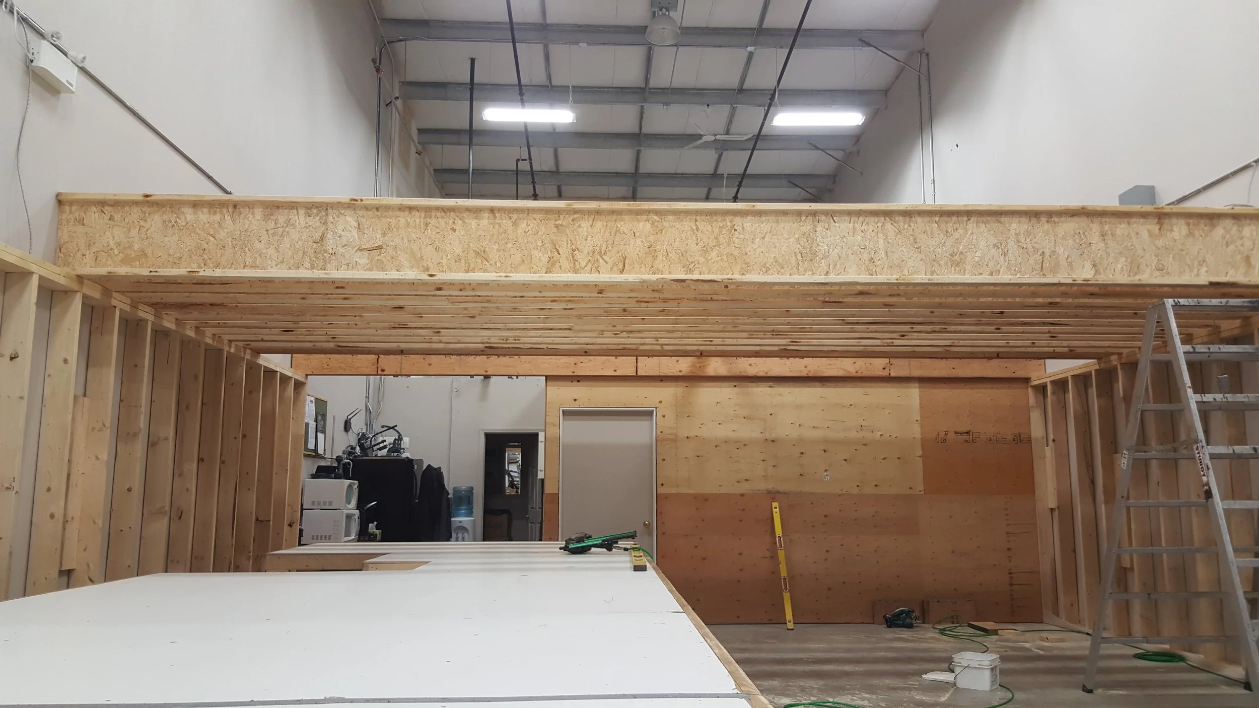 Interior view of a construction site with unfinished wooden framework and an upper loft area. Tools and materials are scattered on the floor, and a ladder is leaning against the right wall.