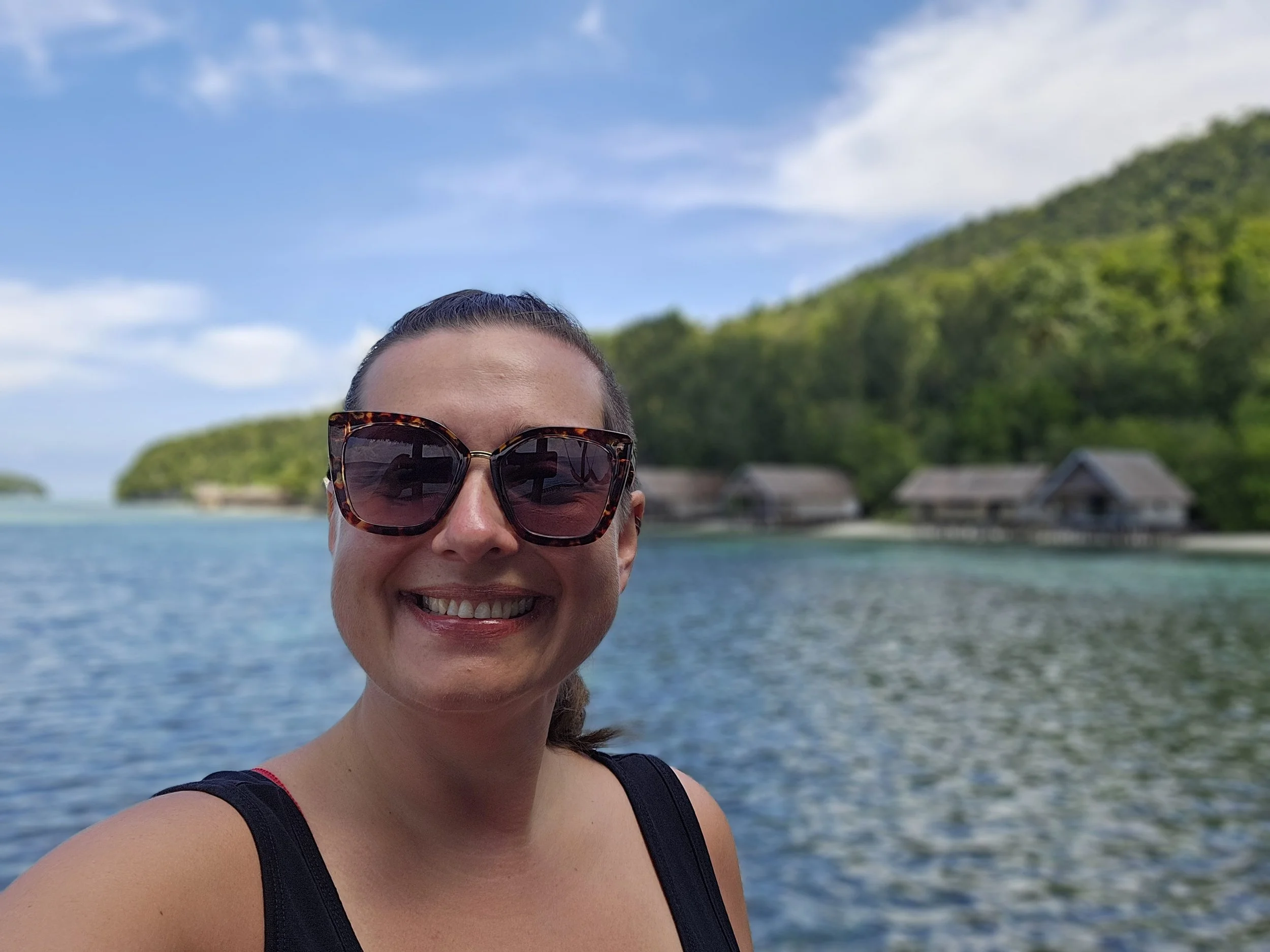 A woman wearing sunglasses smiling in front of a body of water, with green hills and overwater bungalows in the background under a partly cloudy sky.