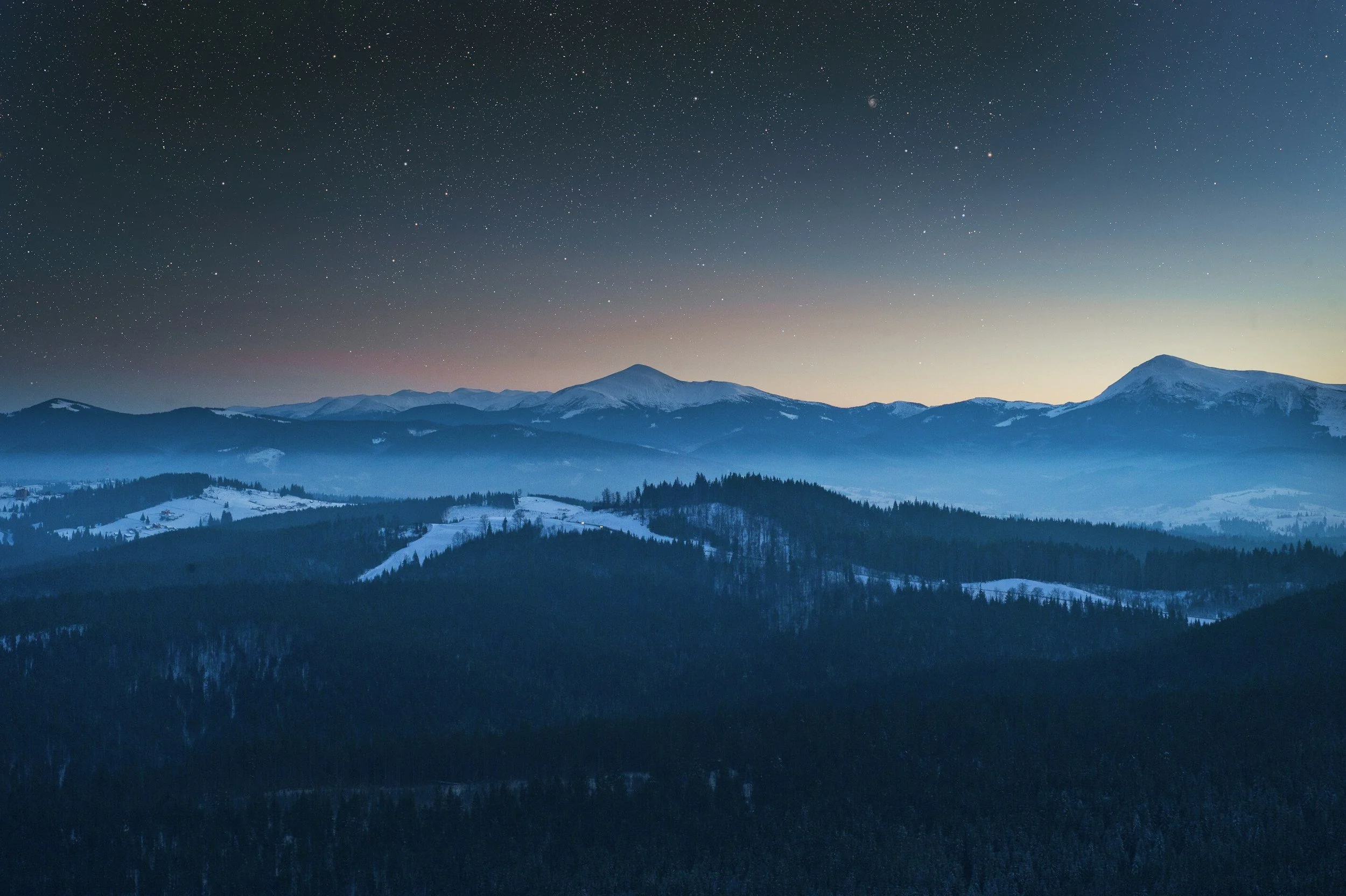 Nighttime view of snow-covered mountain peaks and forested hills under a starry sky with the Milky Way galaxy visible.