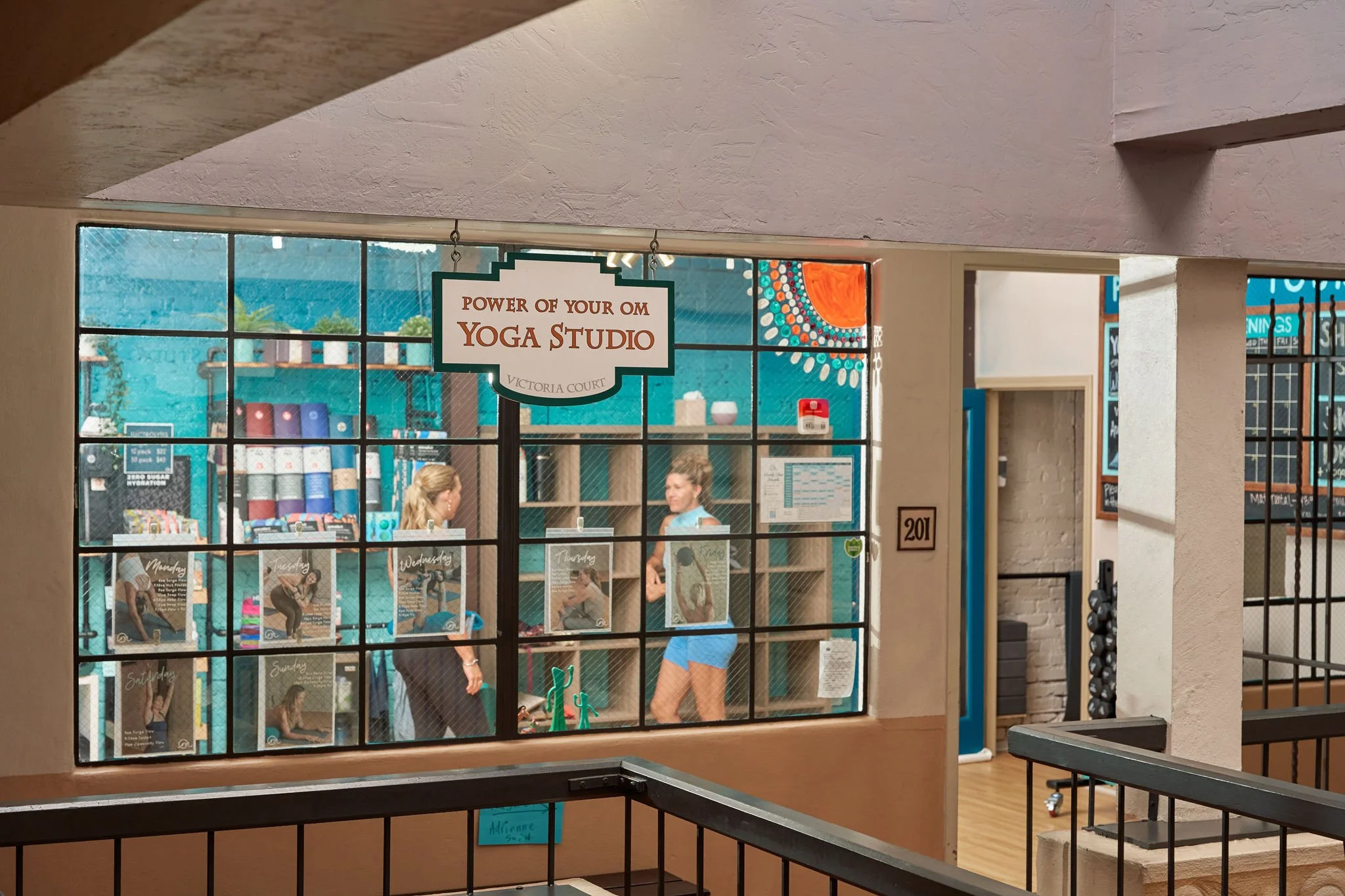 Interior of a yoga studio with a sign that reads 'Power of Your OM Yoga Studio Victoria Court'. The view through the window shows a blue wall, posters of people doing yoga poses, and two women conversing inside.