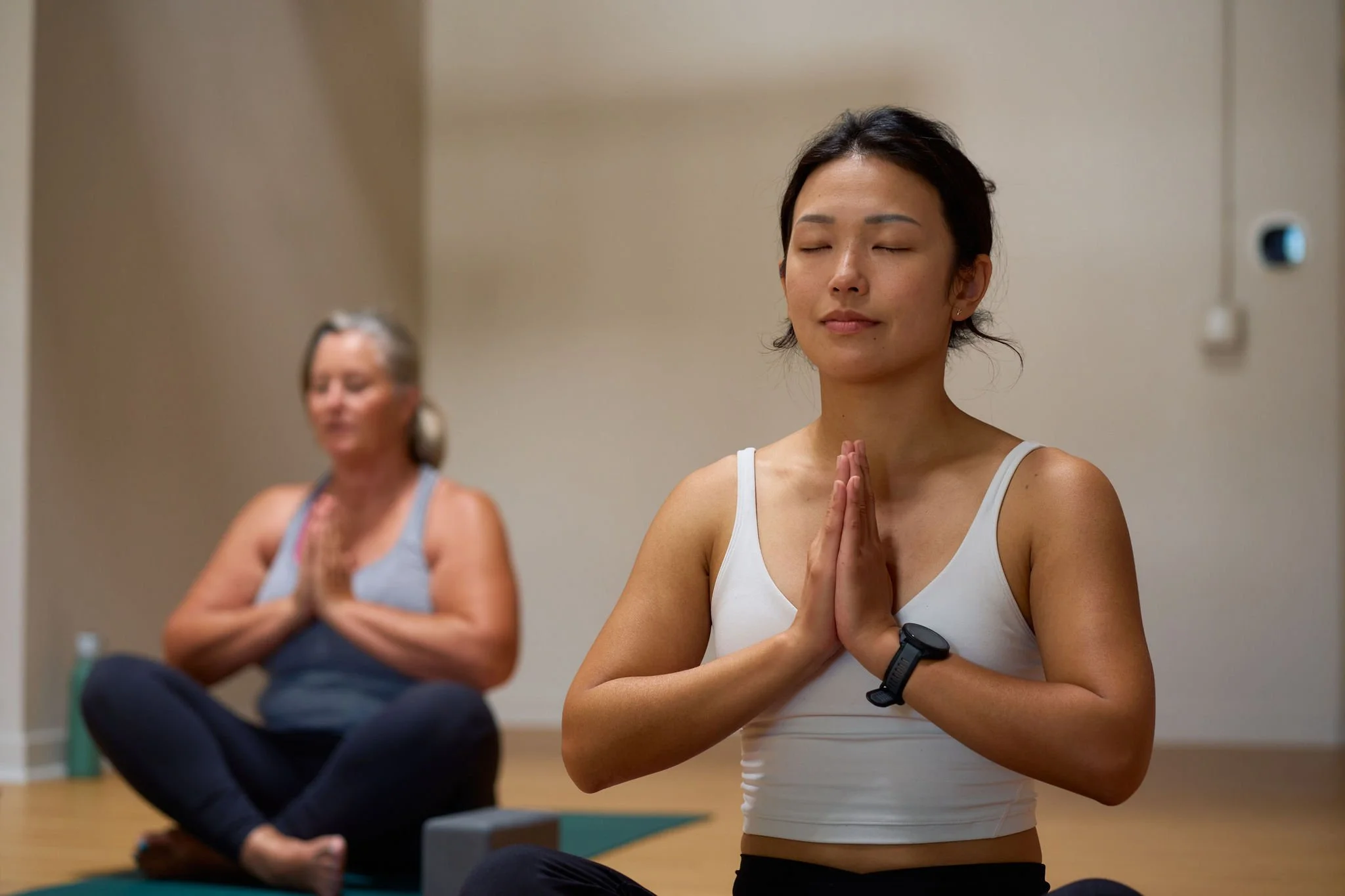 Two women practicing yoga indoors, sitting cross-legged with hands in prayer position and eyes closed.