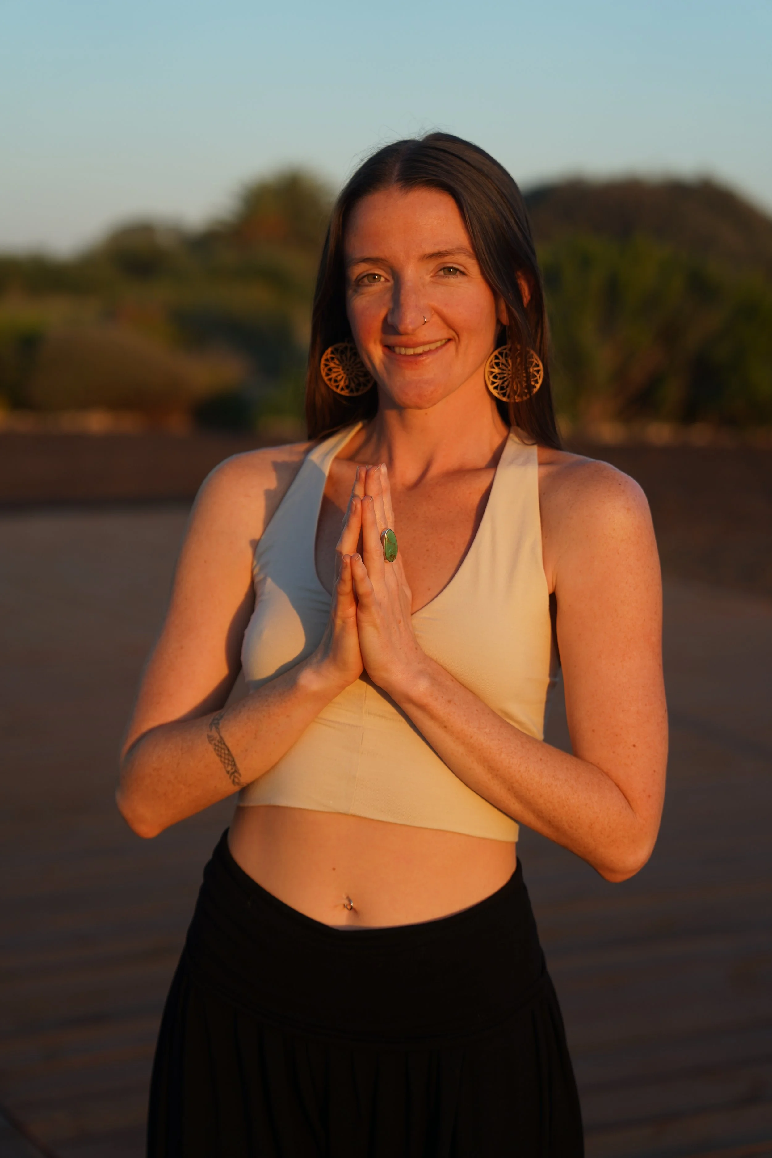A woman standing outdoors during sunset, with her hands pressed together in a prayer pose, wearing a beige crop top, black skirt, and large earrings, with a natural background of trees and sky.