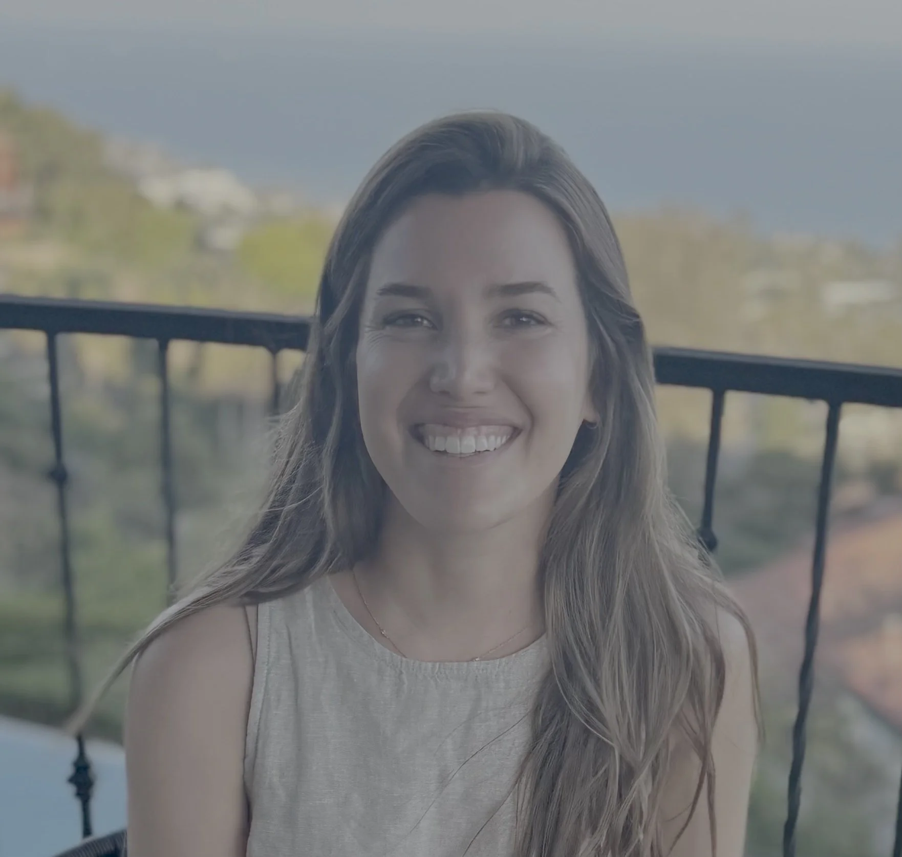 Smiling woman with long, wavy hair wearing a sleeveless top, sitting outdoors with a blurred background of trees and a boat railing.