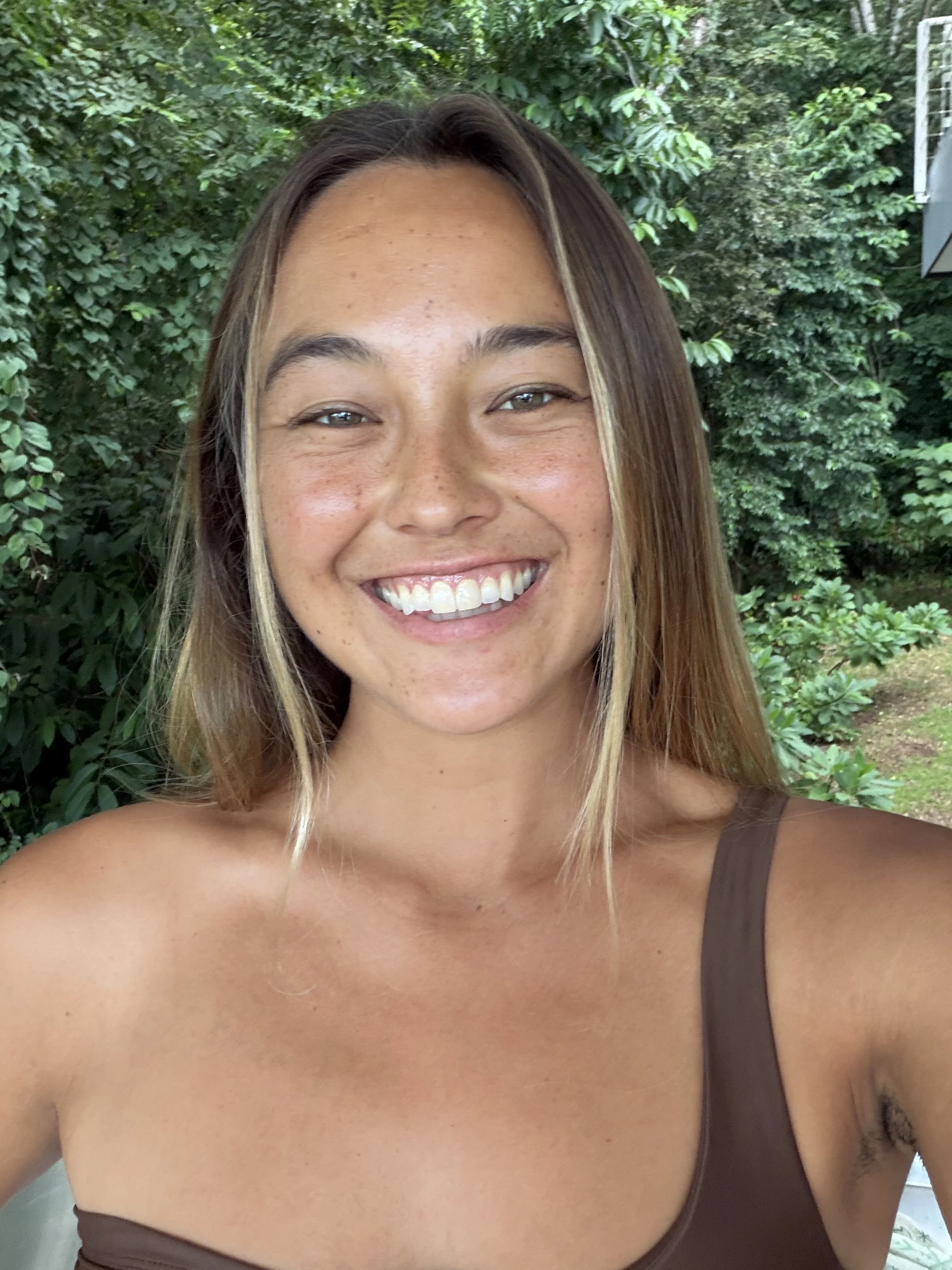 A smiling woman with long, light brown hair and light skin, wearing a brown tank top, standing outdoors in front of greenery.