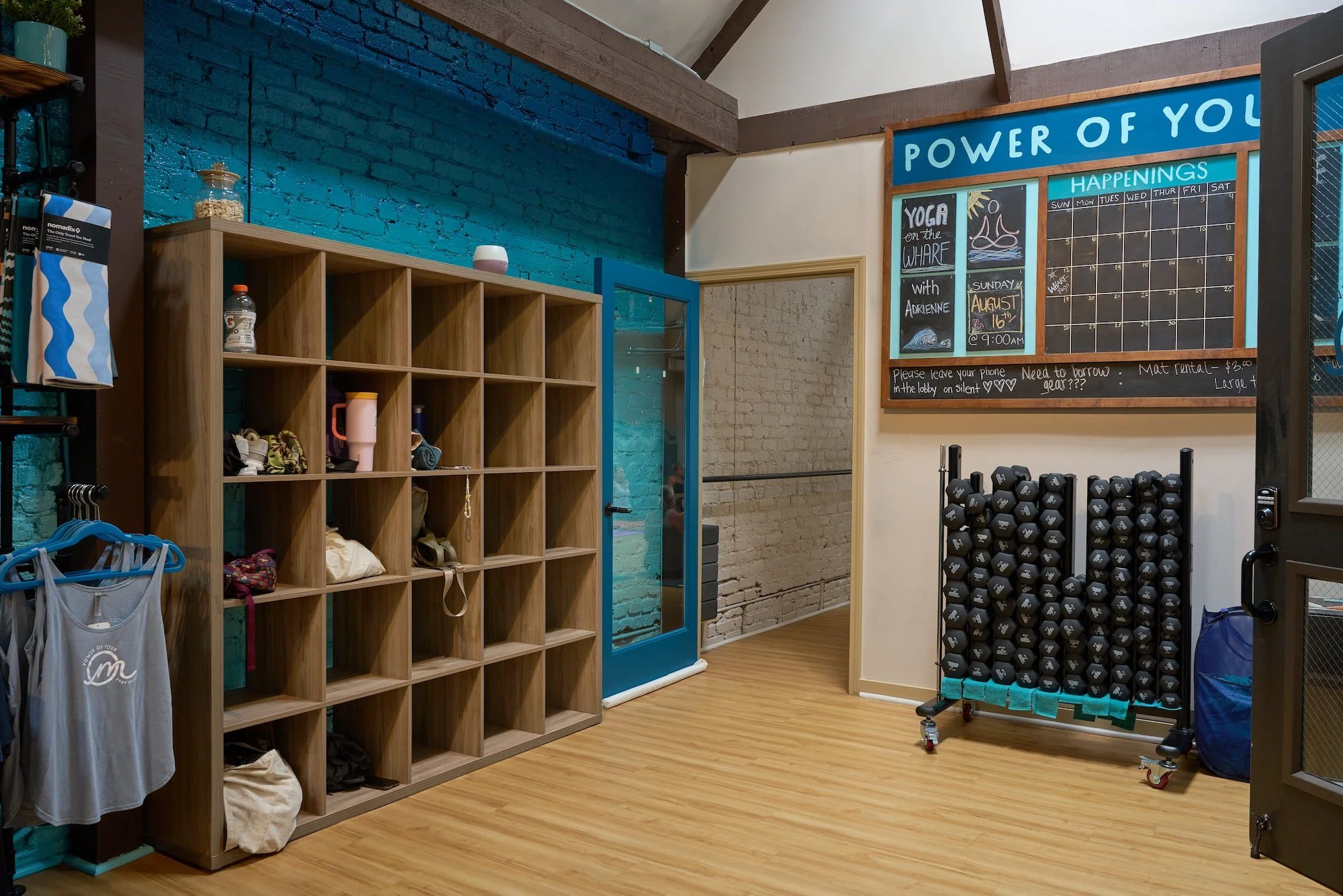 Interior of a fitness or yoga studio with a wooden cubby shelf filled with various items, a blue brick wall, a door with glass, and a large blackboard. The blackboard has a yoga class schedule titled 'Power of You' with handwritten notes about upcomi