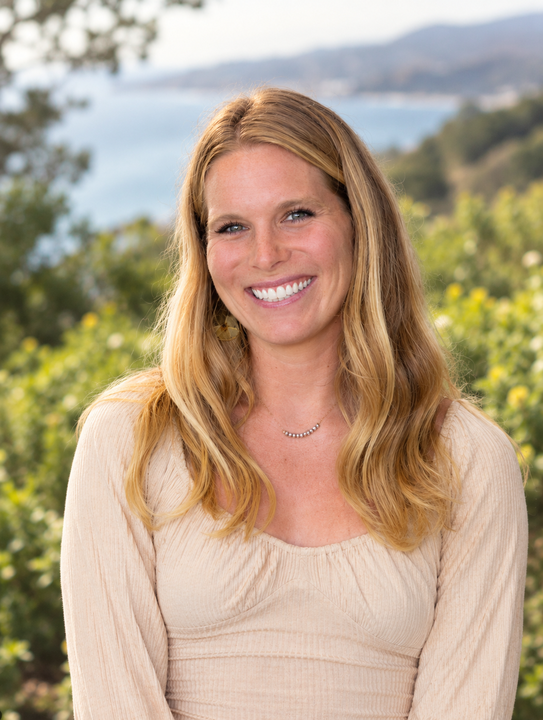 A woman with long, wavy blonde hair smiling outdoors, with a scenic view of water, trees, and hills in the background.