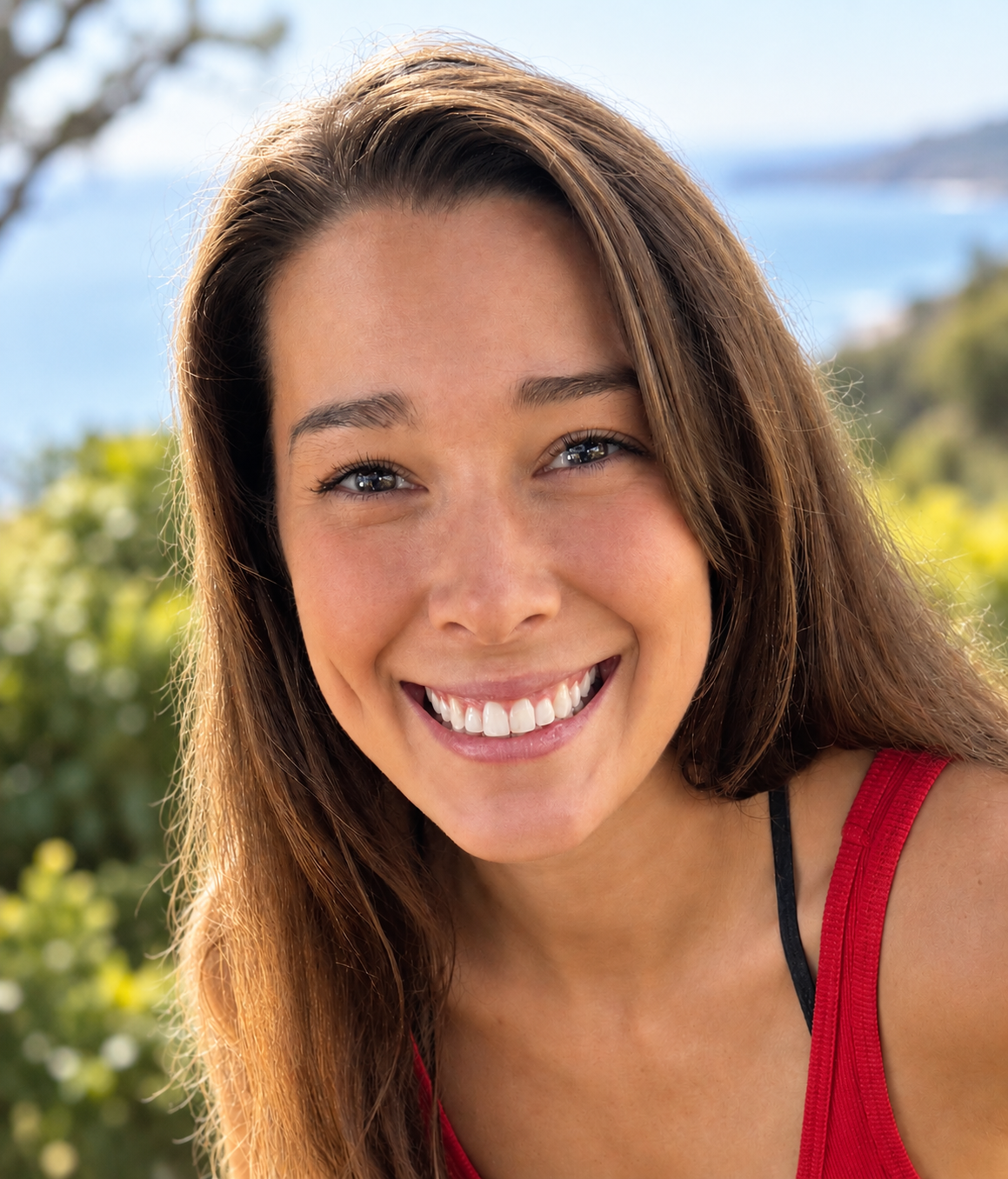 Close-up of a smiling young woman with long brown hair, outdoors near a body of water with greenery in the background.