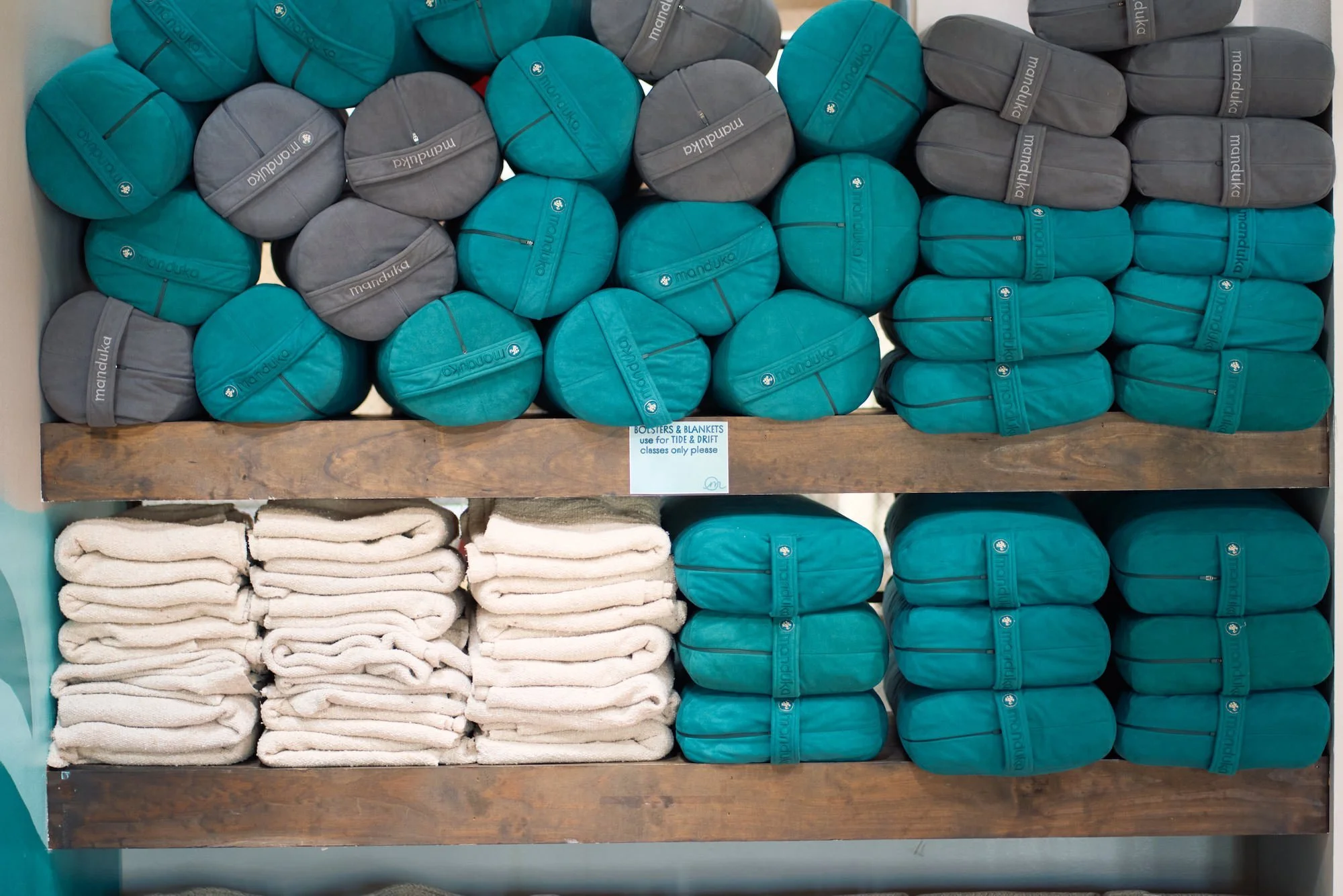Shelves of folded towels and hanging pouches in a store. The pouches are teal and gray with the word 'manduka' printed on them, and the towels are white. A small sign indicates the pouches are for tide and drift classes only.