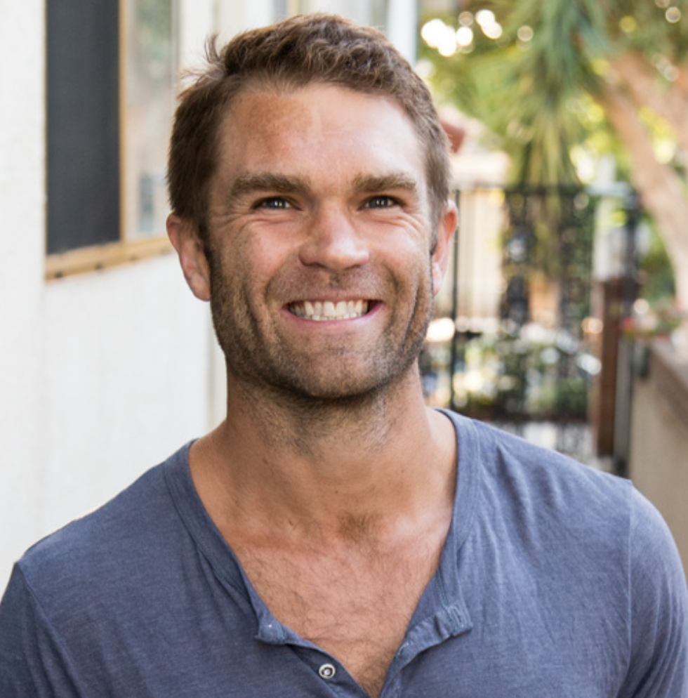 A man smiling outdoors with trees and a building in the background