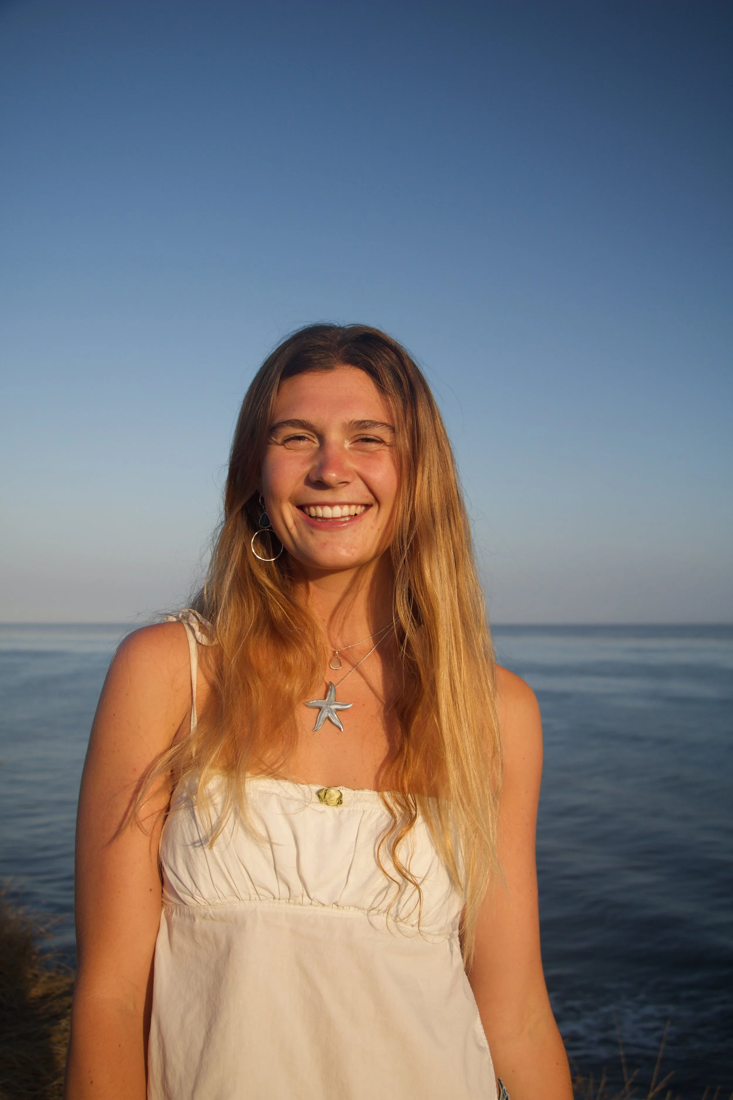A young woman with long, wavy, blond hair smiling at the camera on a beach with calm water and a clear sky in the background.