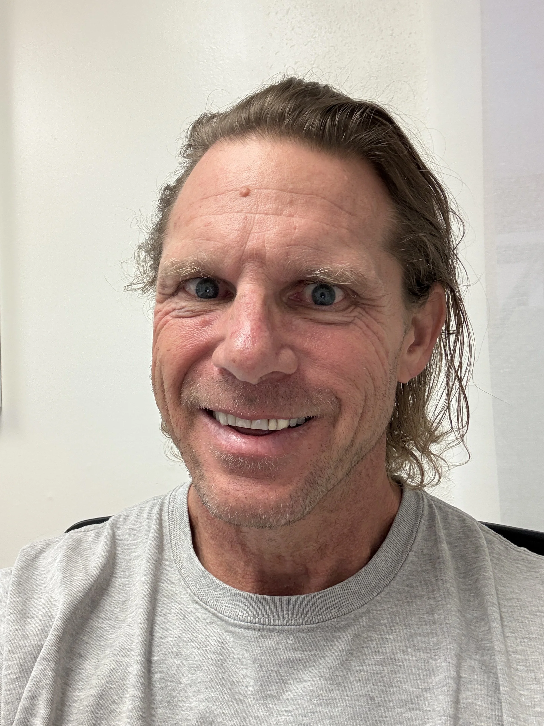 A close-up of a middle-aged man with blue eyes, smiling, with long, slightly curly brown hair, wearing a light gray shirt, standing against a plain light-colored wall.