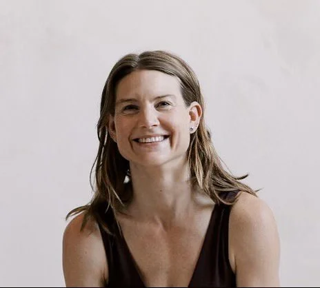 A smiling woman with shoulder-length brown hair, wearing a sleeveless black top, in front of a plain light-colored background.