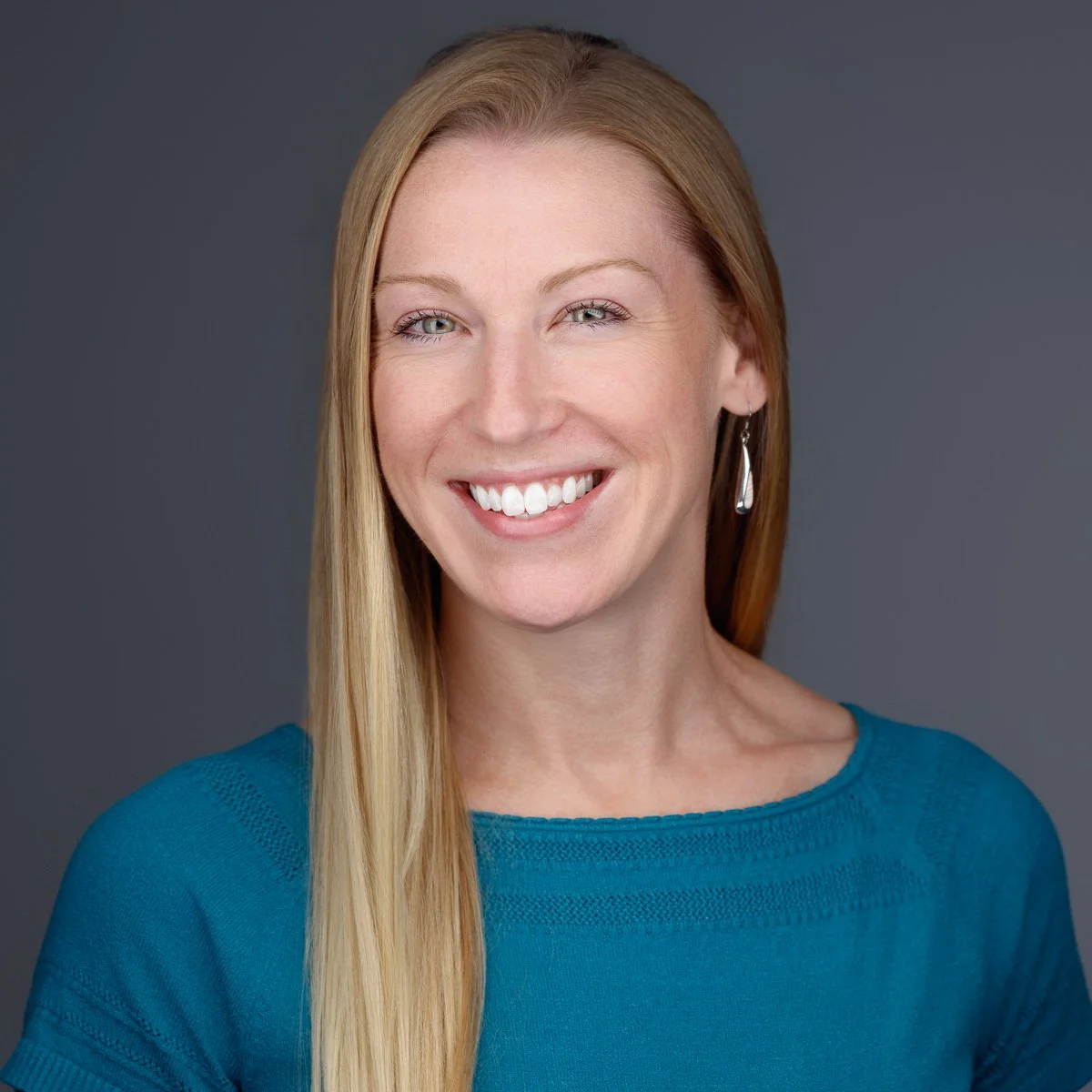 A woman with long blonde hair, wearing a blue top and dangling earrings, smiling against a gray background.