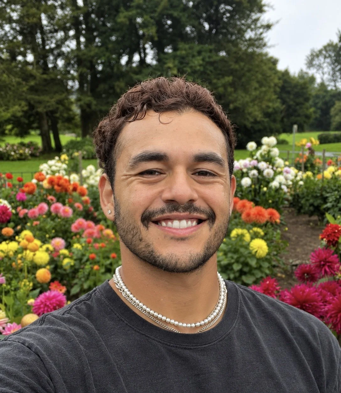 A man smiling in front of a colorful flower garden with various blooming flowers and trees in the background.