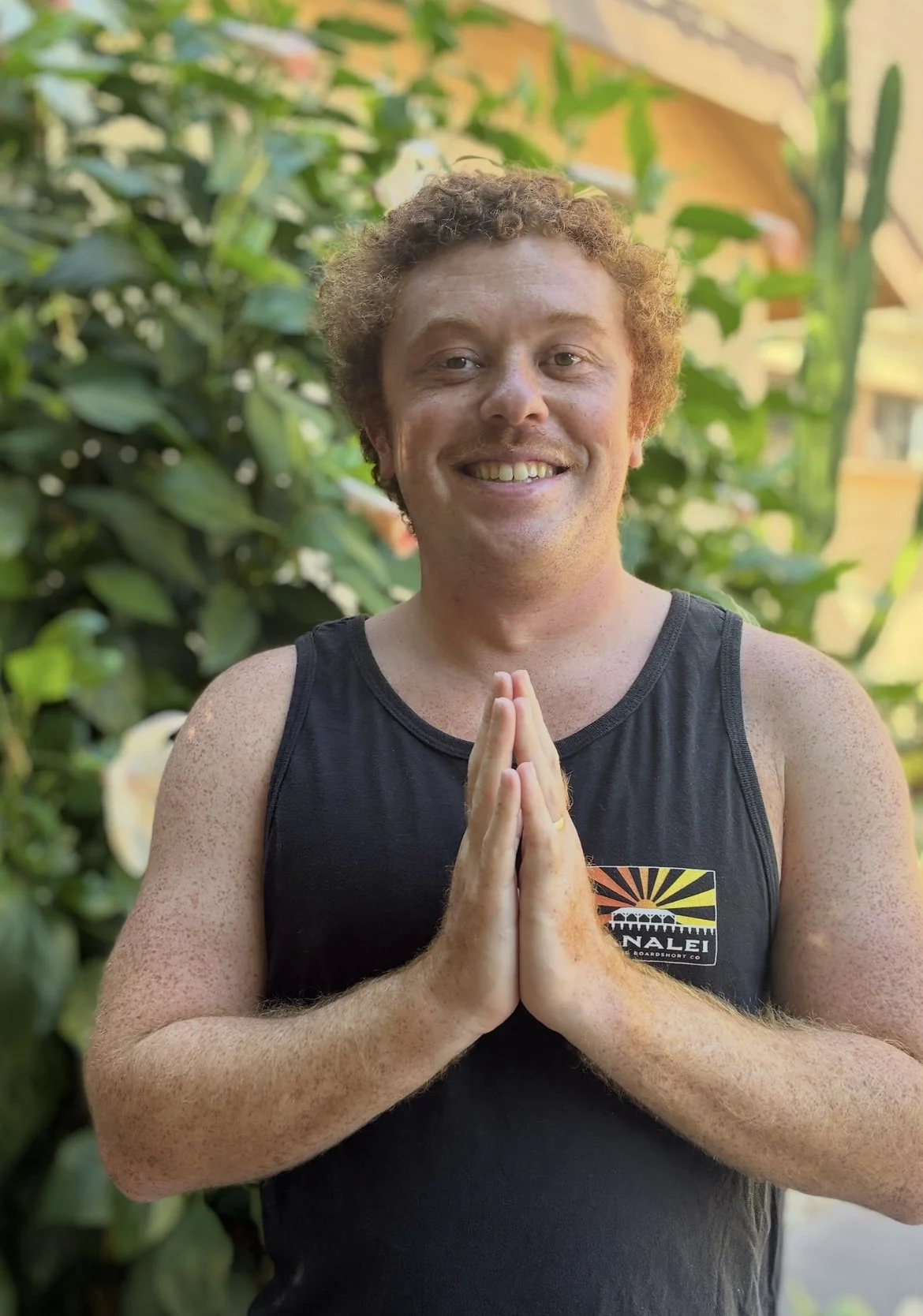 A smiling man with curly hair and freckles wearing a black tank top, standing outdoors with green foliage in the background, holding his hands together in a prayer pose.
