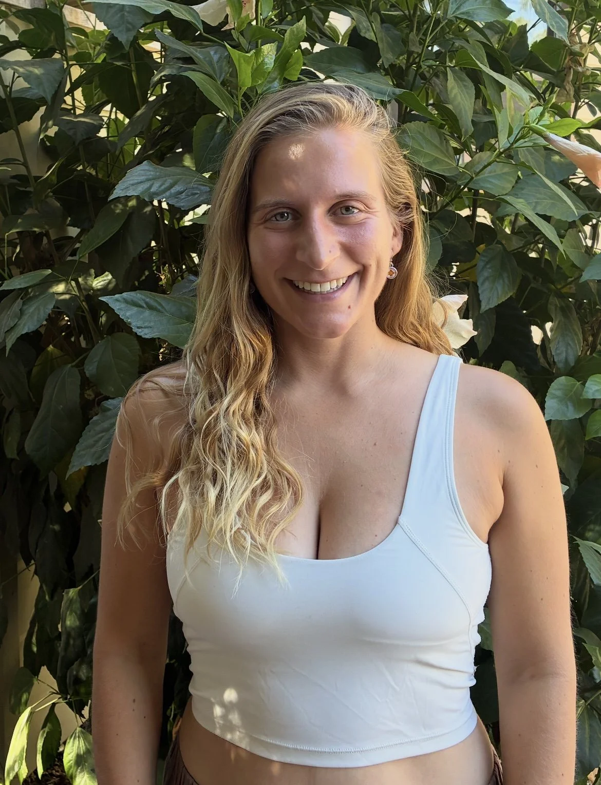 A young woman with long blonde curly hair smiling in front of green leafy plants, wearing a white sleeveless top.